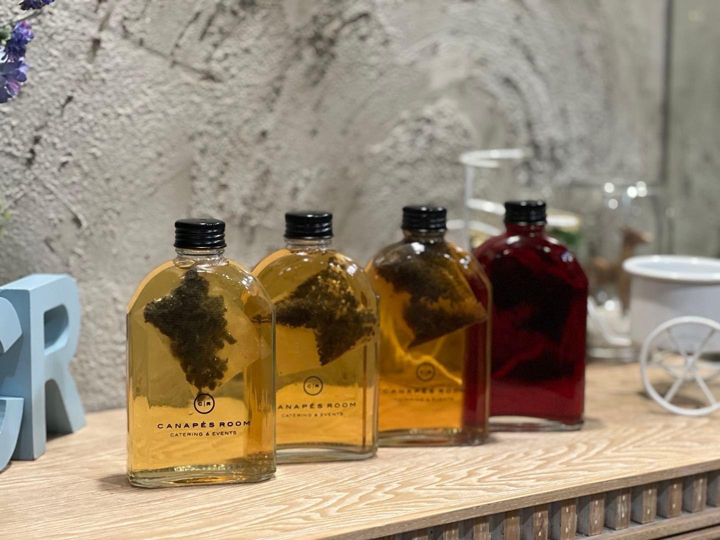 Four bottled drinks on a shelf. Three are golden, one is red, with tea leaves visible.