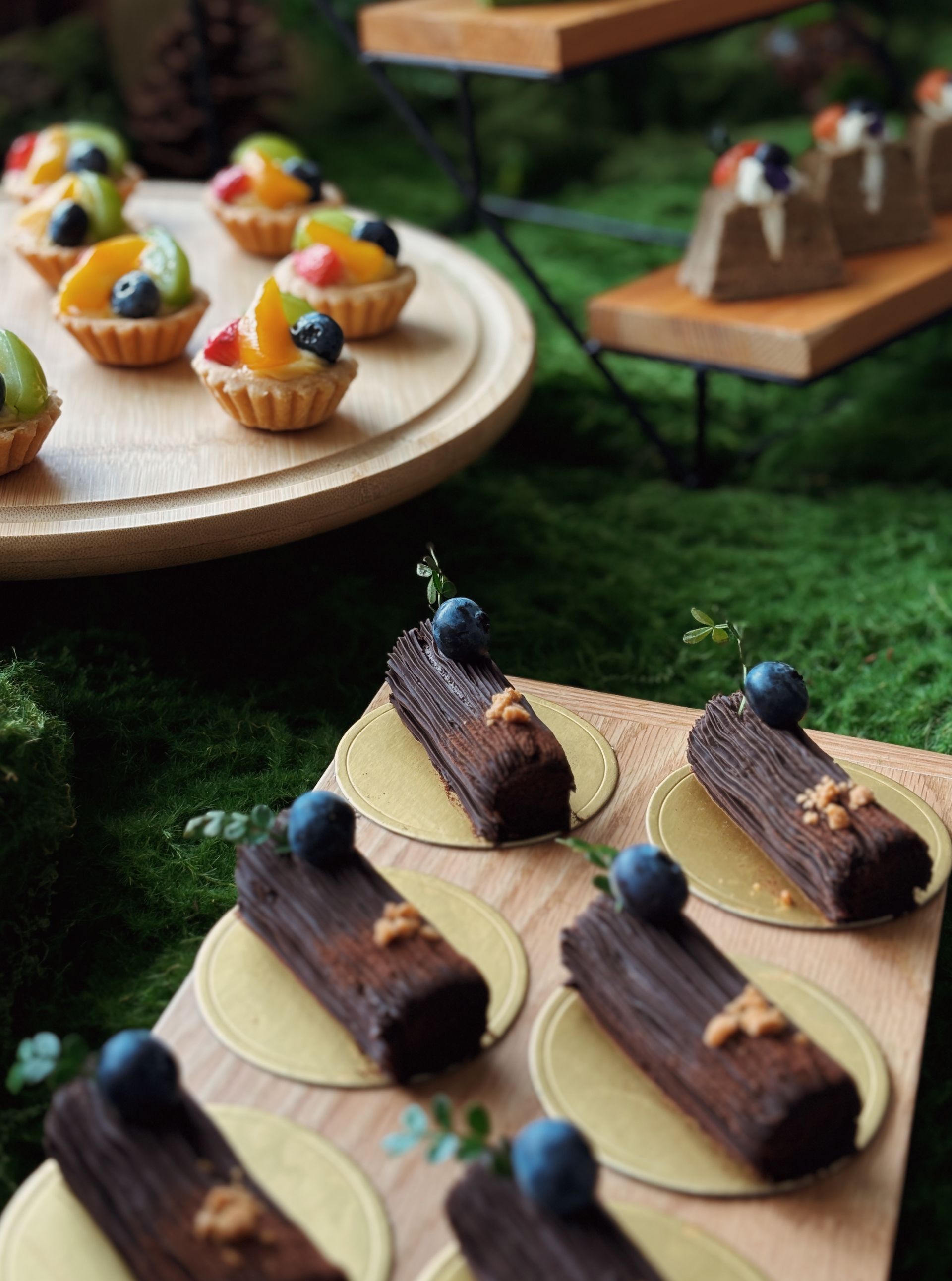 Mini fruit tarts and chocolate log desserts on wooden platters, set on moss.