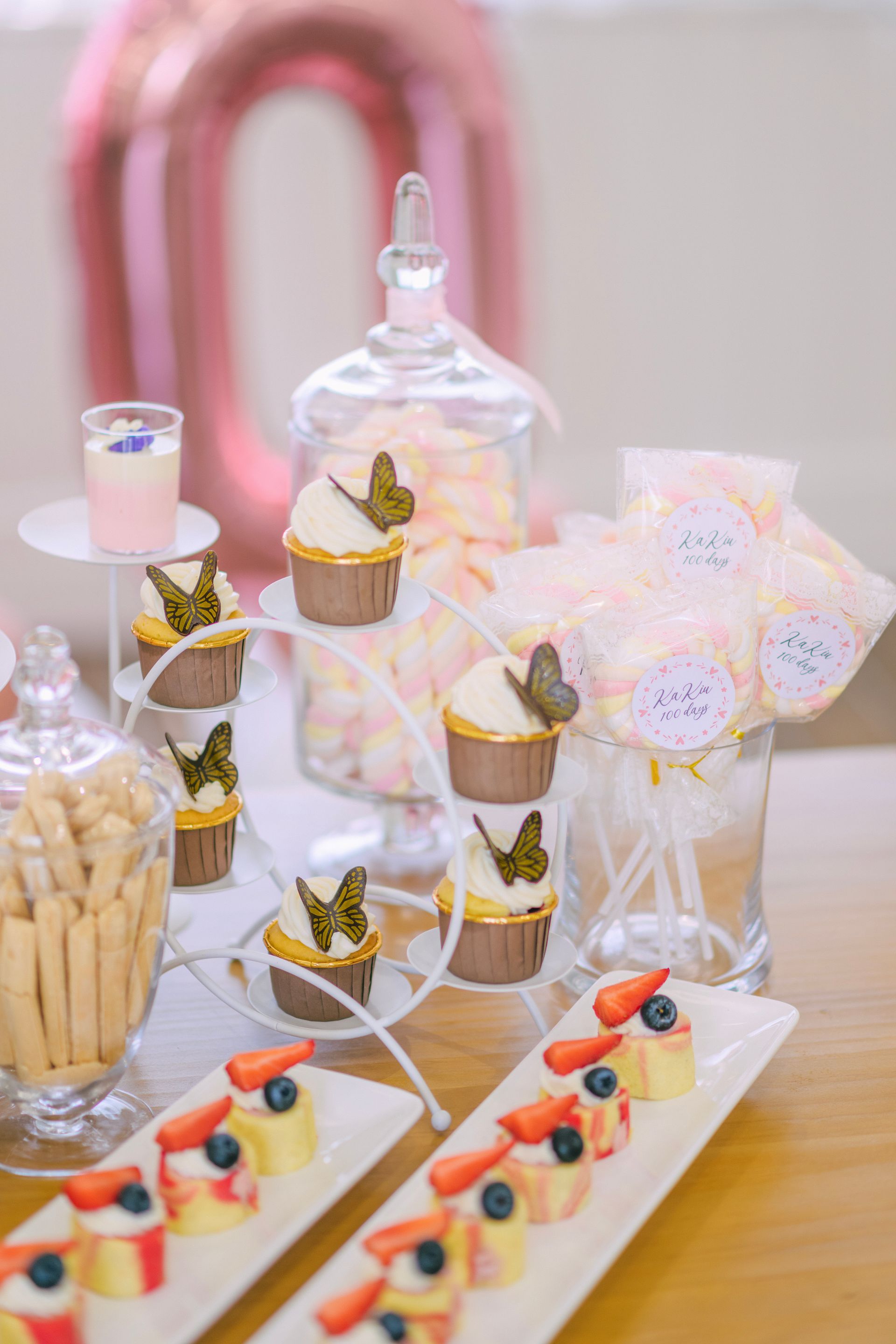 Dessert table: cupcakes with butterfly toppers, cake bites with fruit, sweets in jars, and a pink balloon.