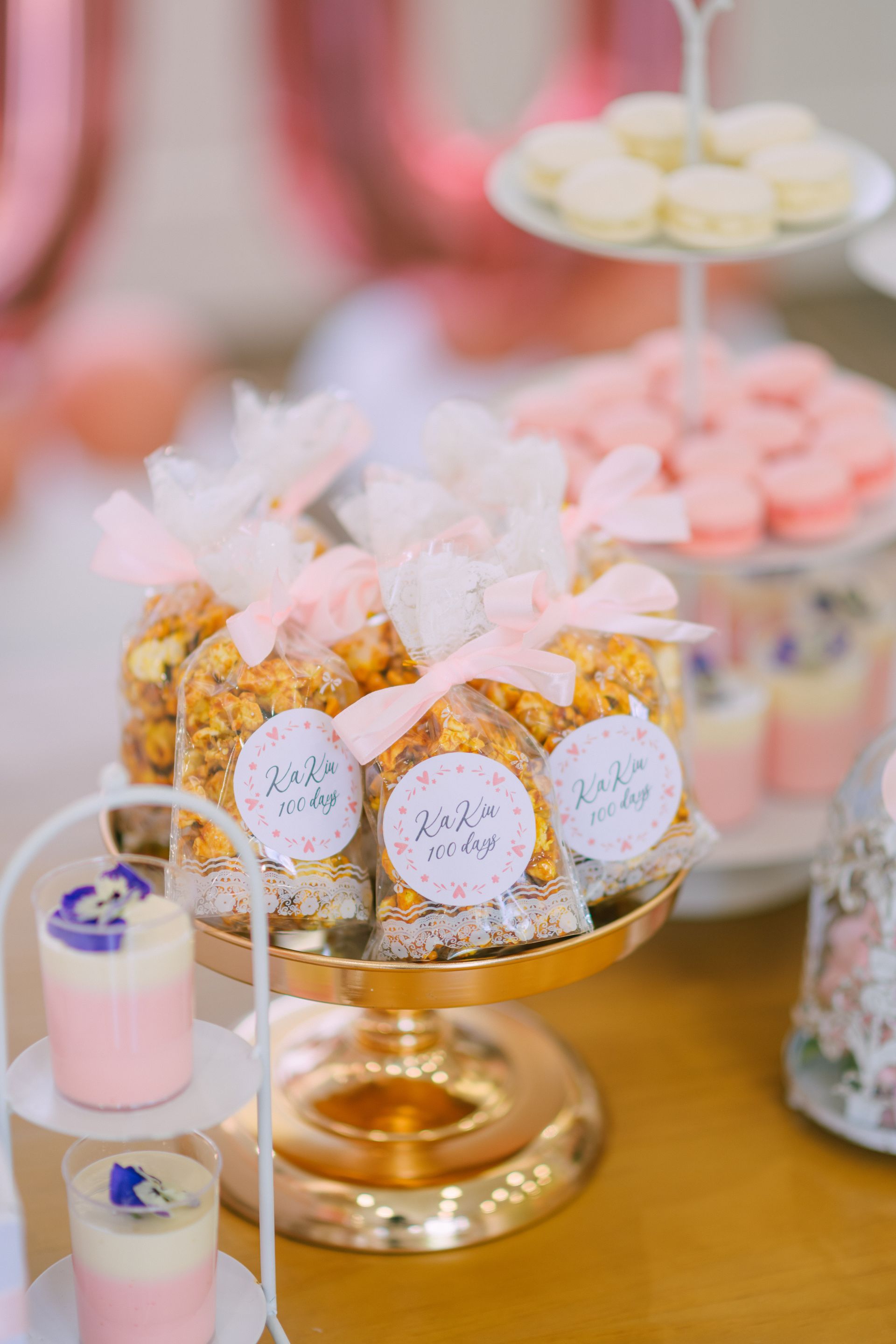 Treats on a gold stand: popcorn bags with pink ribbons, macarons, and layered desserts.