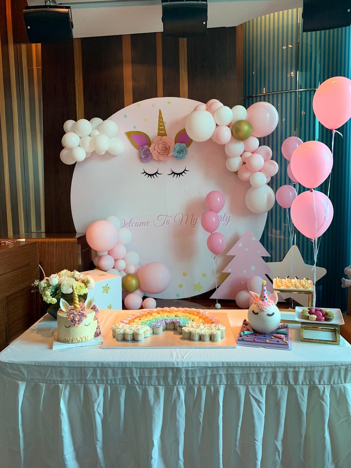 Unicorn-themed party setup with cake, cupcakes, balloons, and decorations in pink and white.