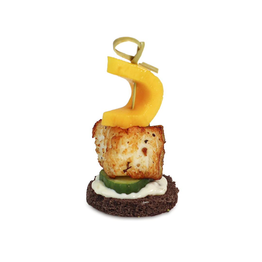 Appetizer skewer: black bread, cucumber, fried cheese, mango slice, secured by a toothpick.