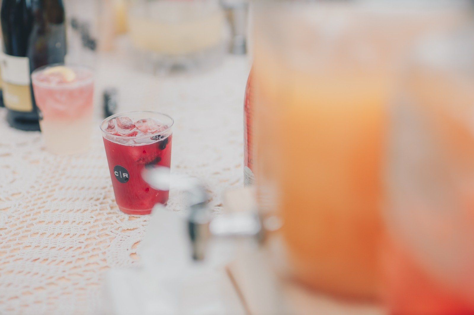 A glass of red cocktail with ice and berries sits on a white lace tablecloth near a juice dispenser.