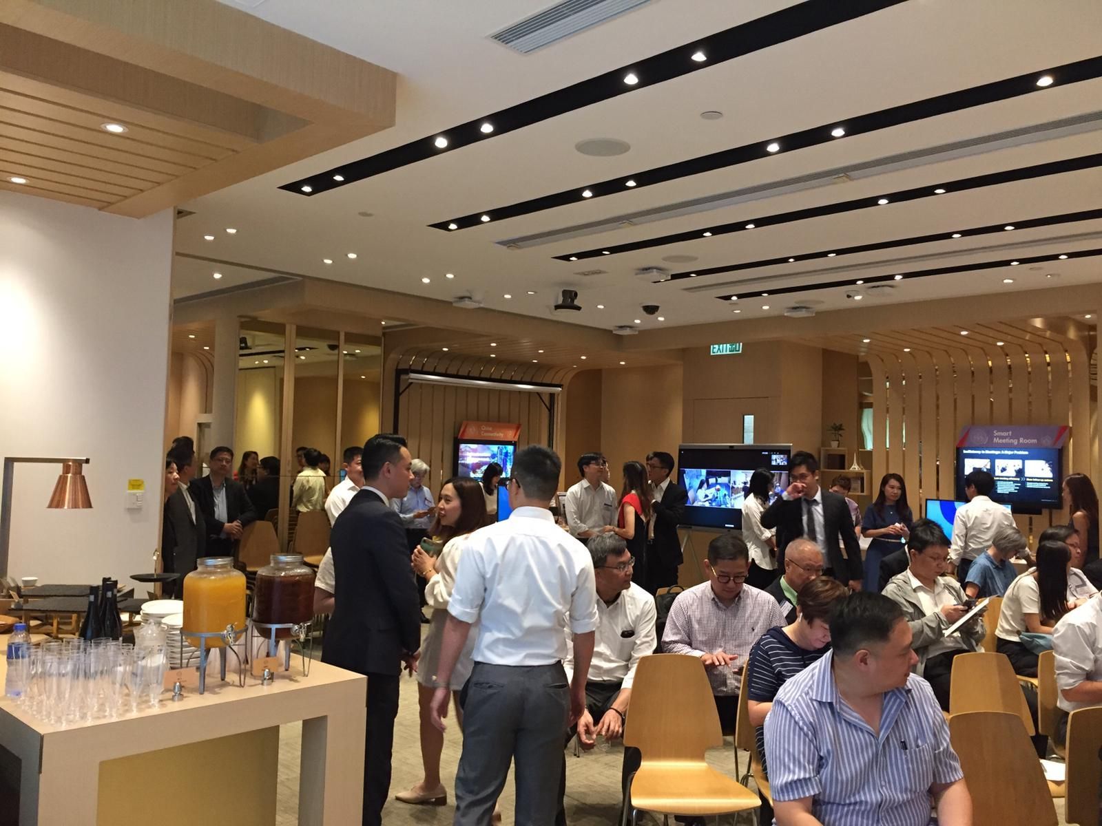People gather in a modern room, likely at a business event. Tables, screens, and refreshments visible.
