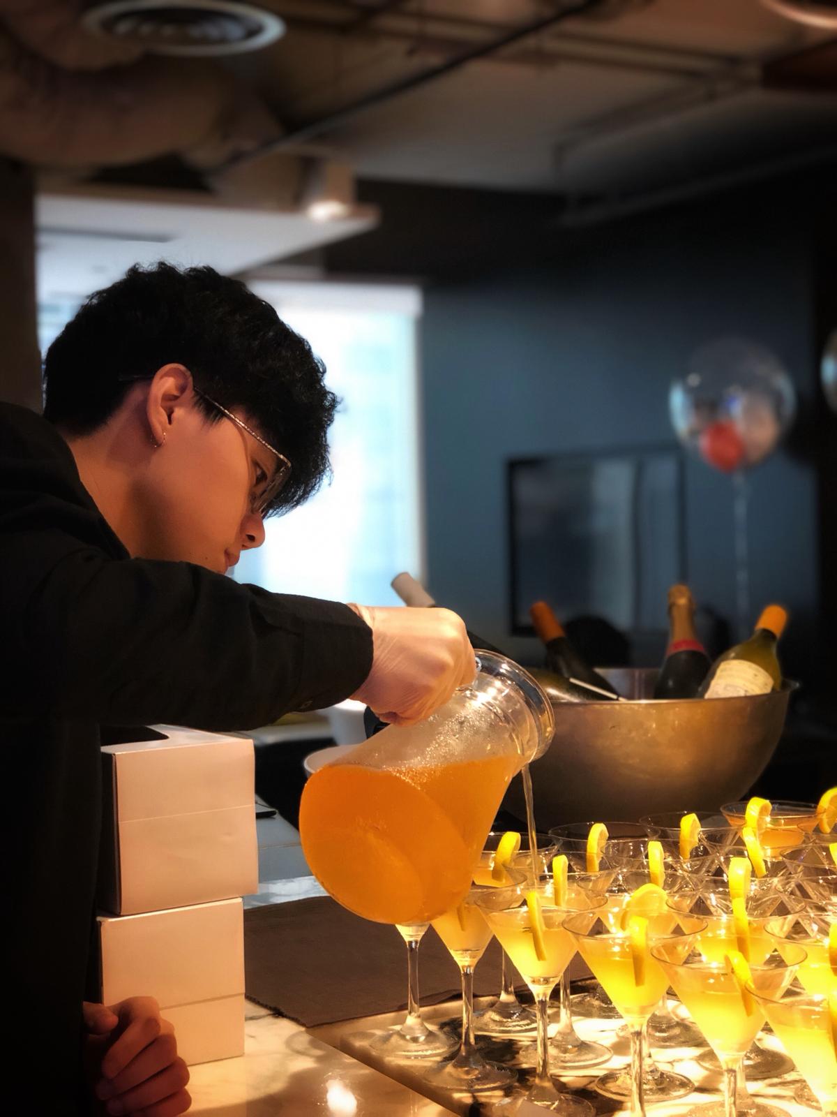 Bartender pouring orange liquid from a pitcher into rows of cocktail glasses at a bar.