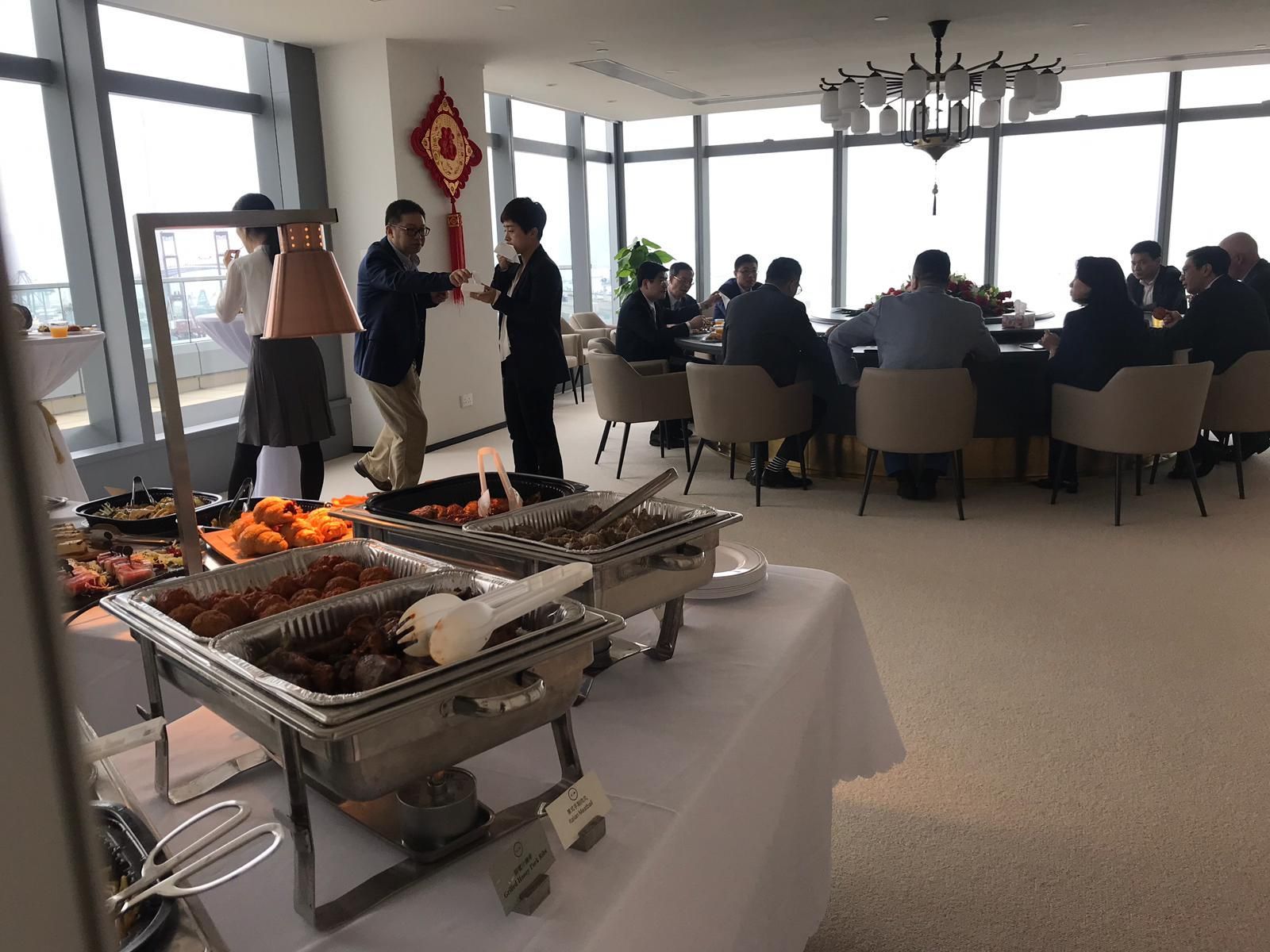 Buffet table with food; people at tables and standing, large windows in a room.
