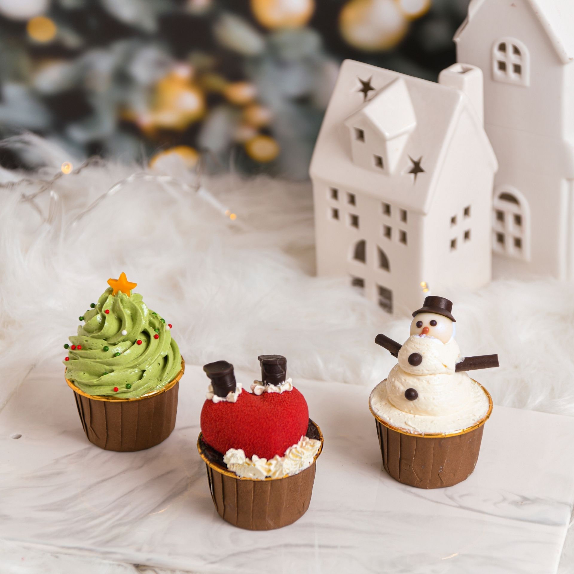 Christmas cupcakes decorated as a tree, Santa, and snowman, with white houses in the background.