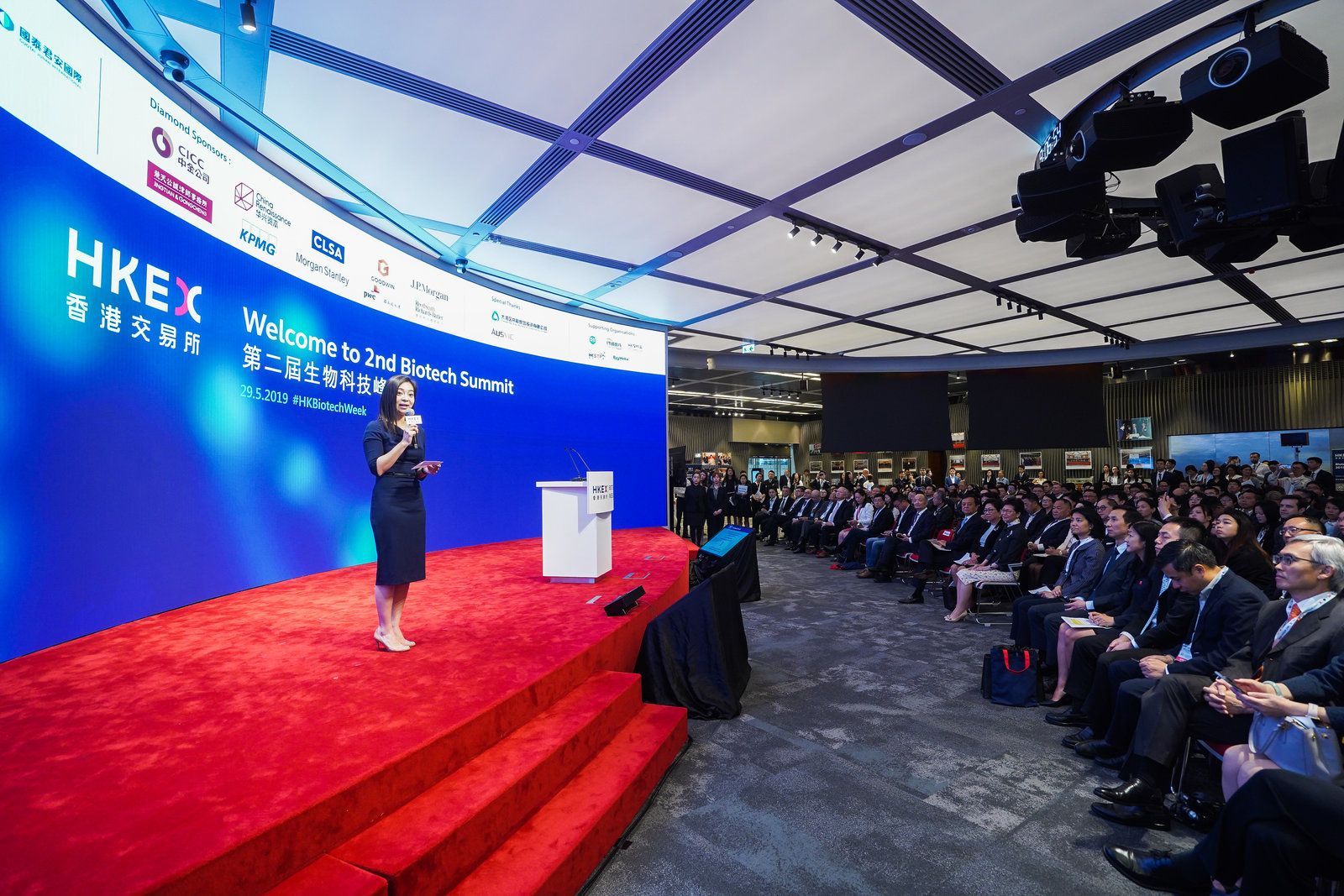 Woman speaking at a conference on a red carpet stage with a blue backdrop that says