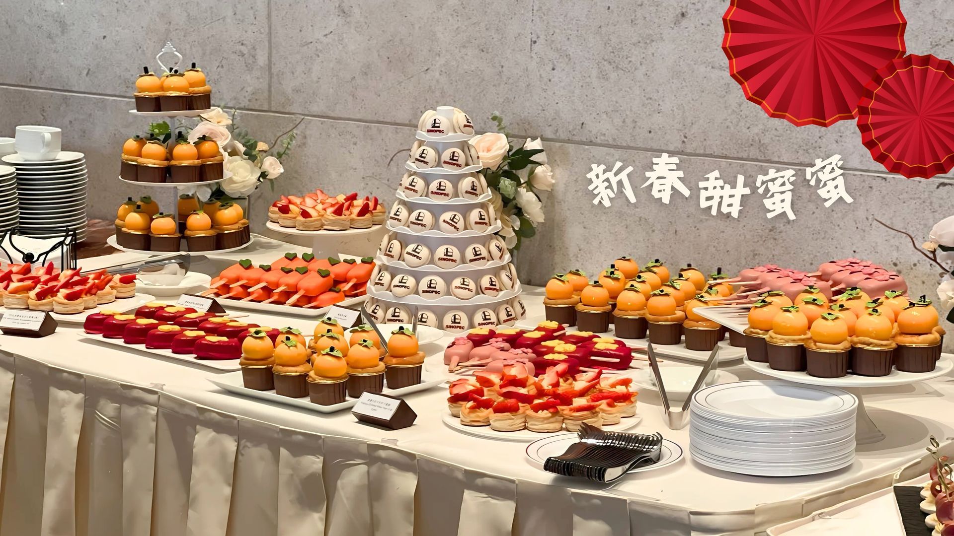 甜點桌上擺放著各種甜點，包括紙杯蛋糕、糕點和多層蛋糕。上方懸掛著紅色紙扇和橫幅。