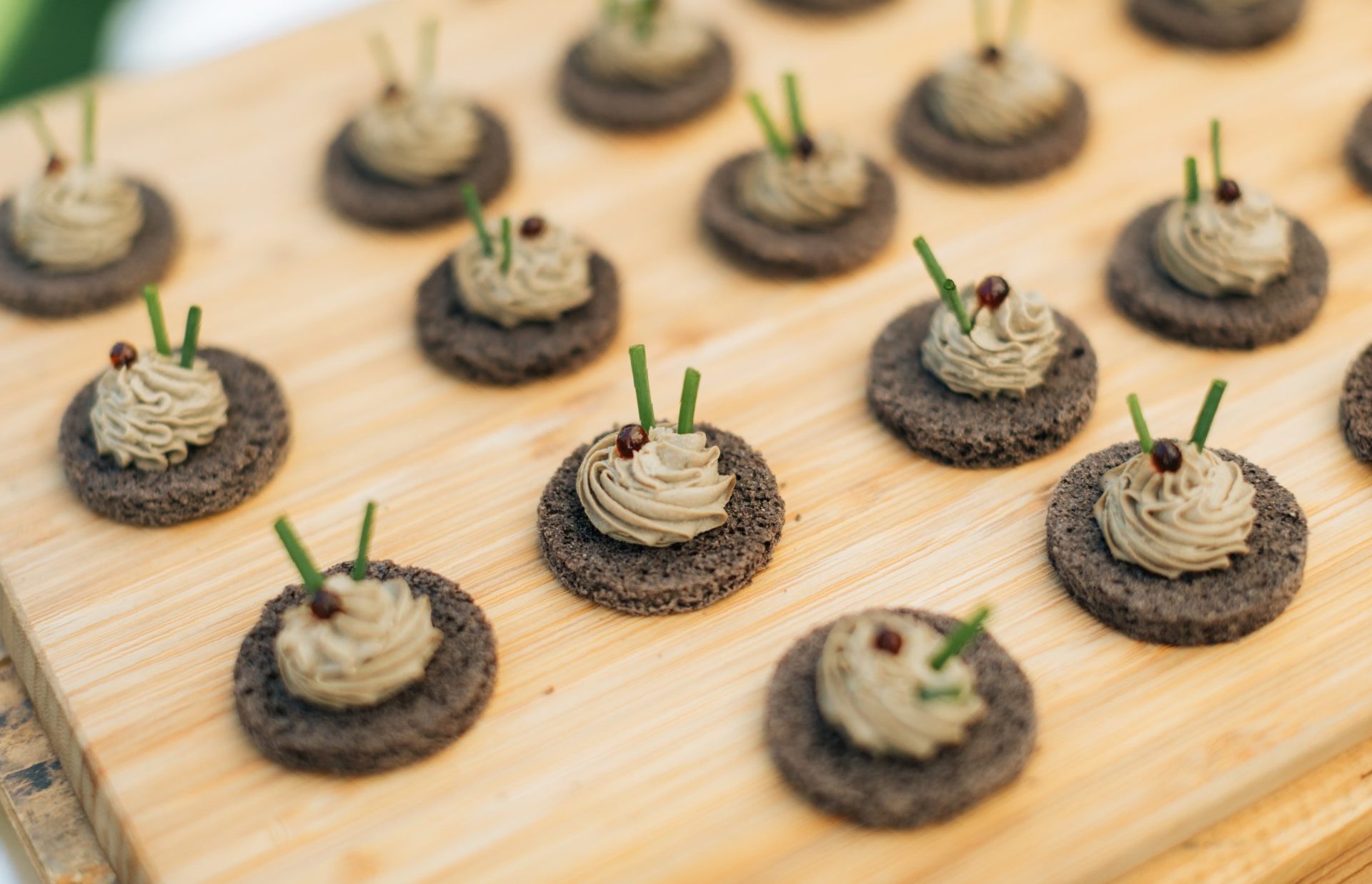 Appetizers: Dark circular crackers topped with tan mousse, chives, and red peppercorns, on a wooden board.