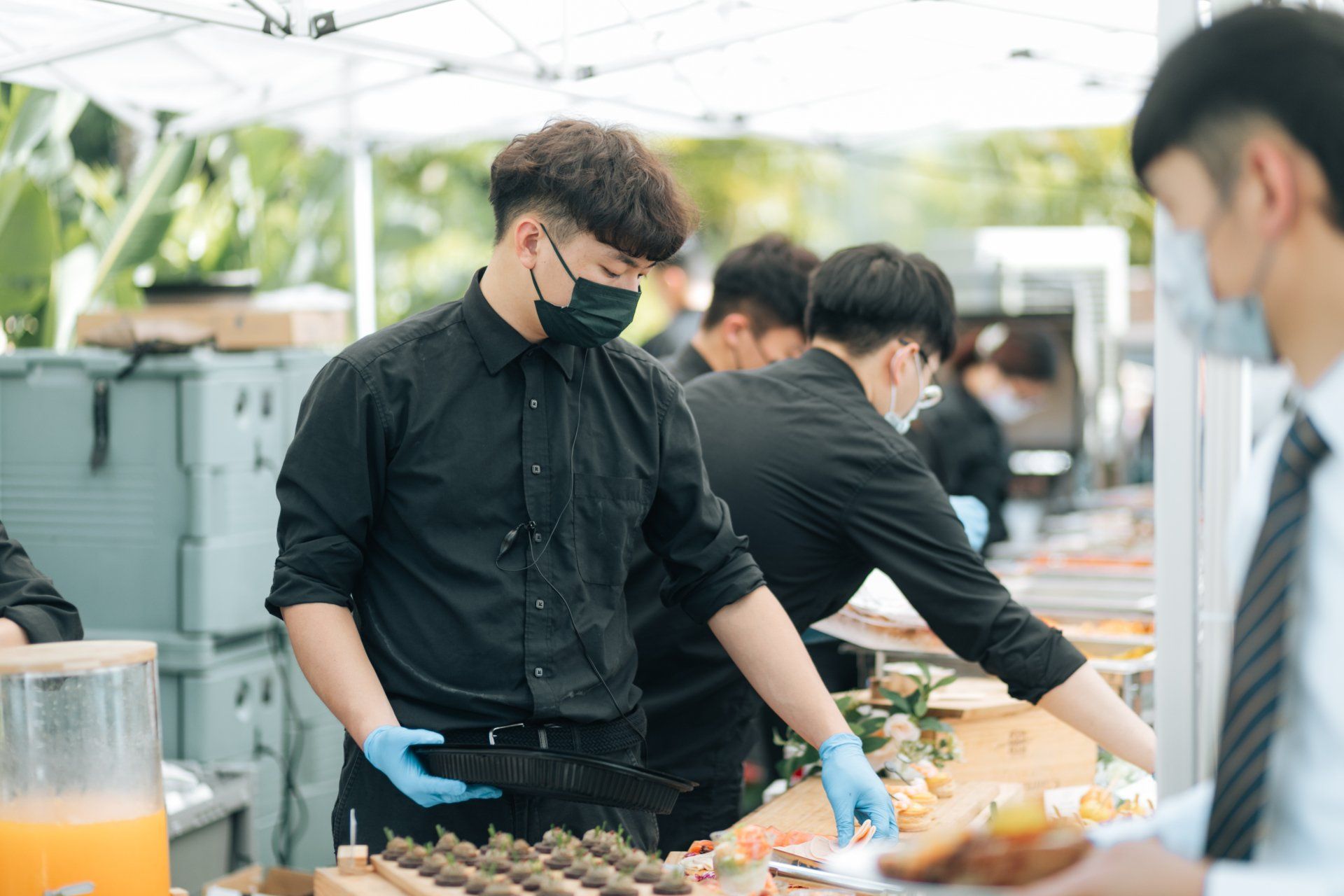 Catering staff wearing masks and gloves serve food at an outdoor event.