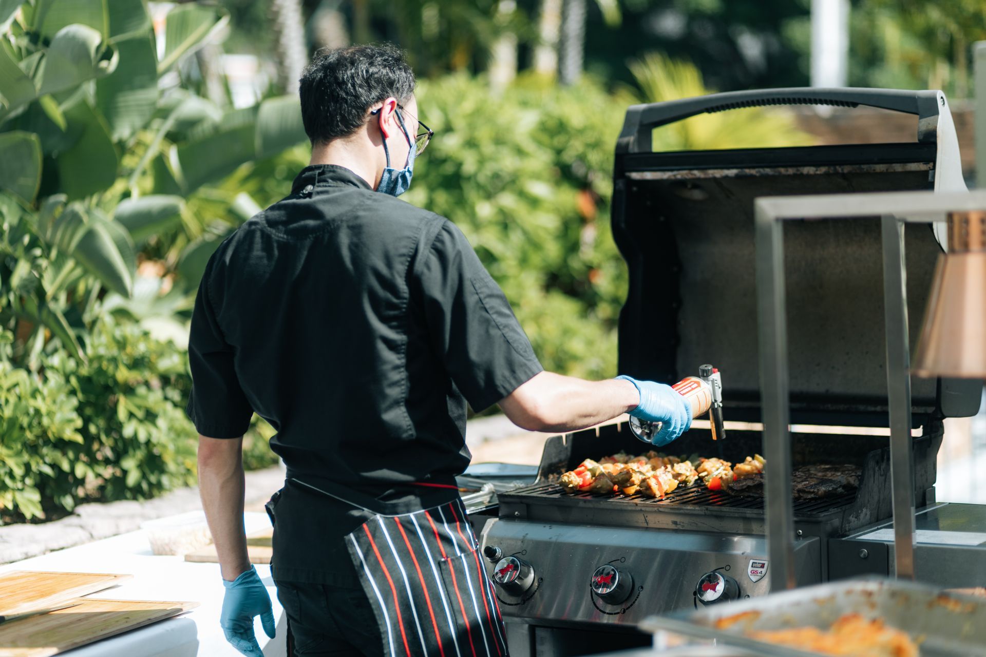 Chef grilling food outdoors, wearing a mask and gloves, near a buffet table.