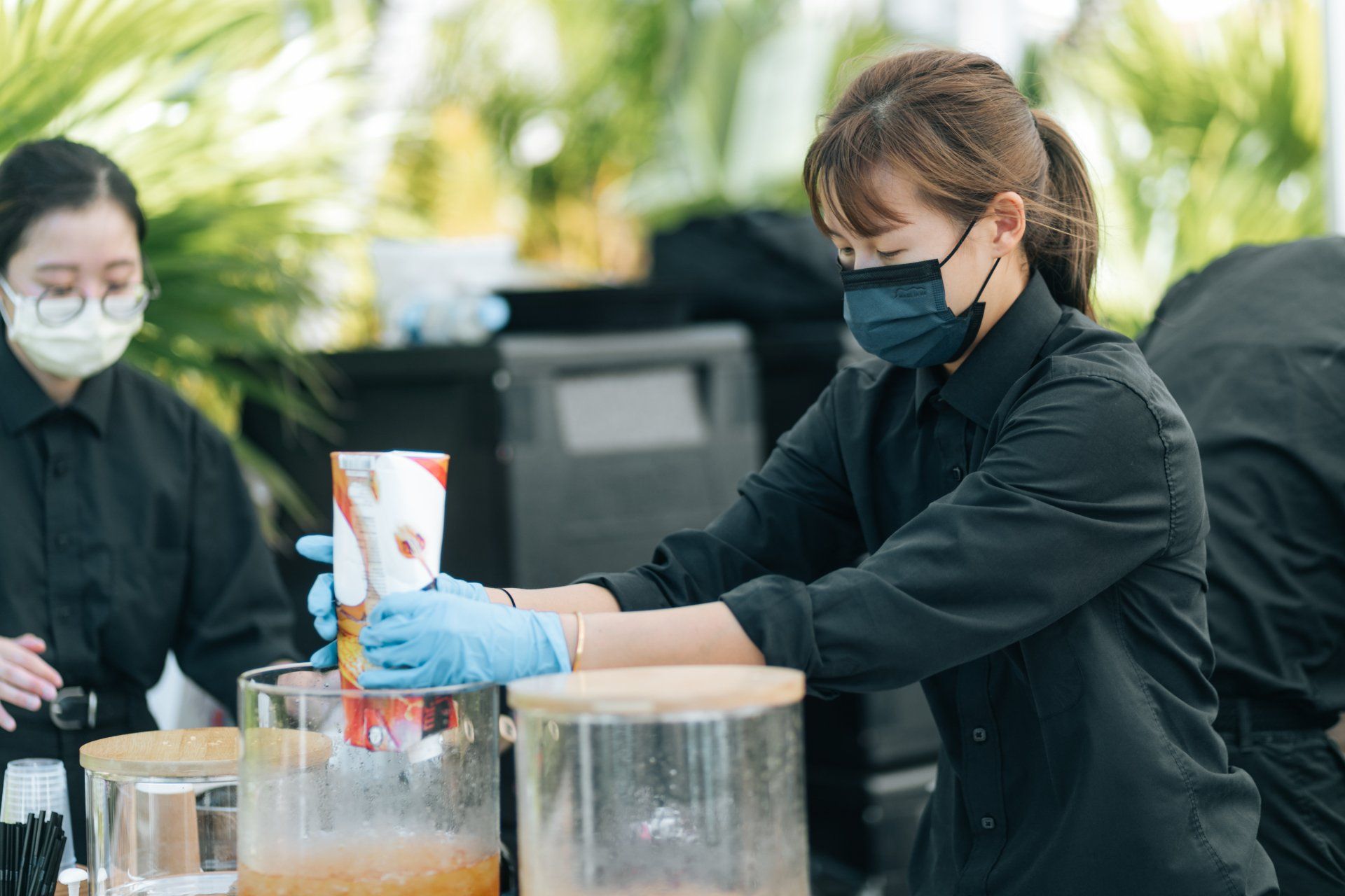 Two people wearing masks and gloves at a drink station. One is pouring from a container.