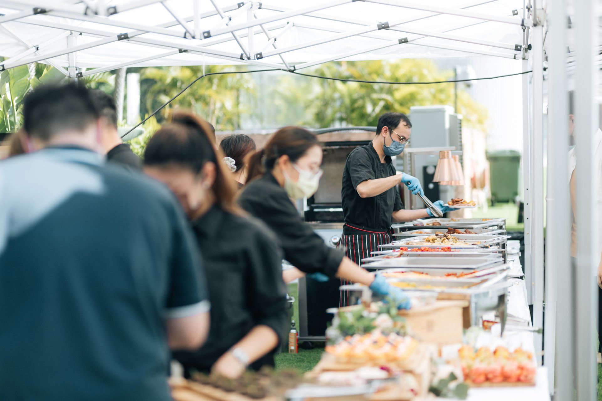 People at a catered outdoor event, serving food from buffet tables under a canopy. Some wear masks.