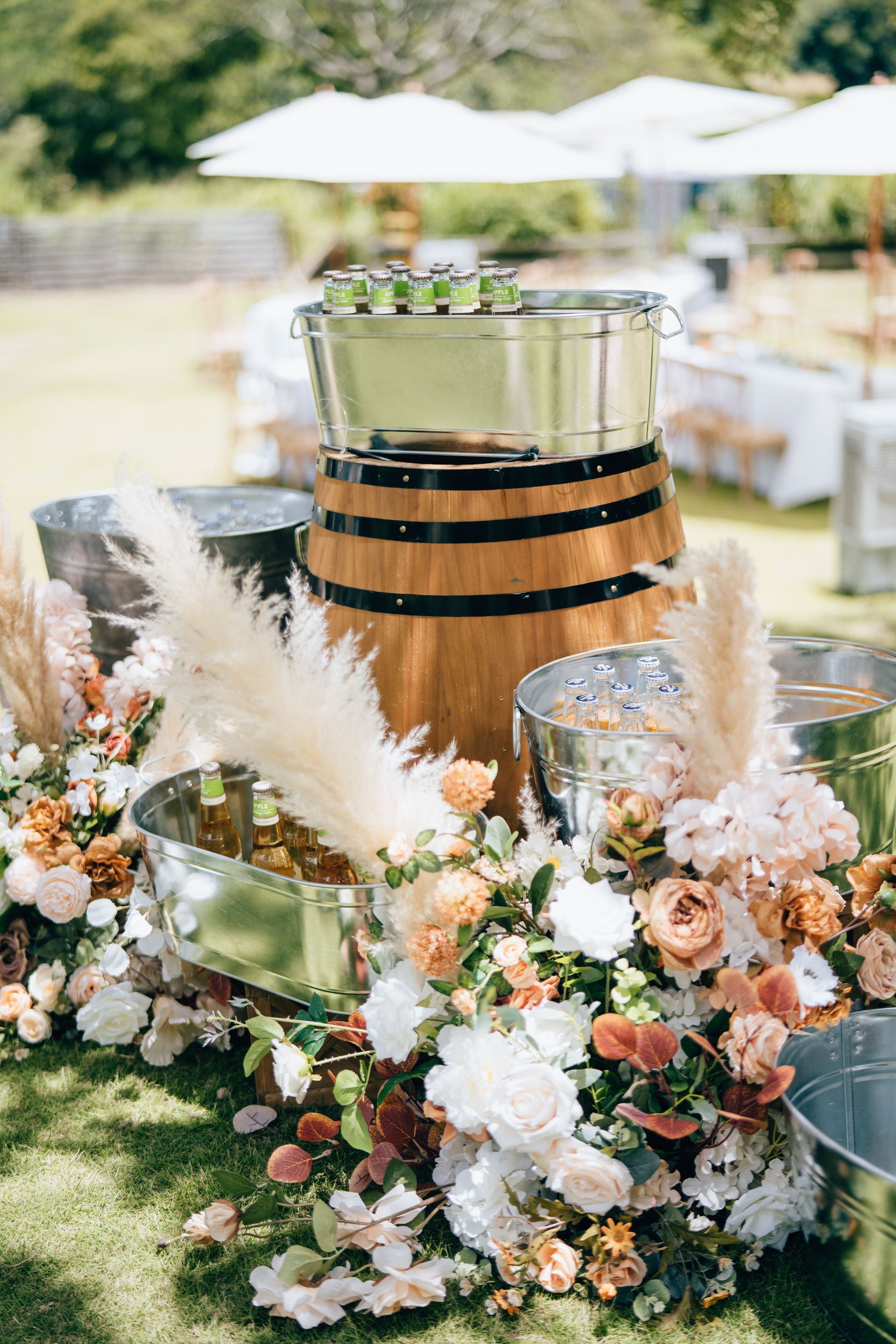 Outdoor beverage station with a barrel, metal buckets, flowers, and drinks.