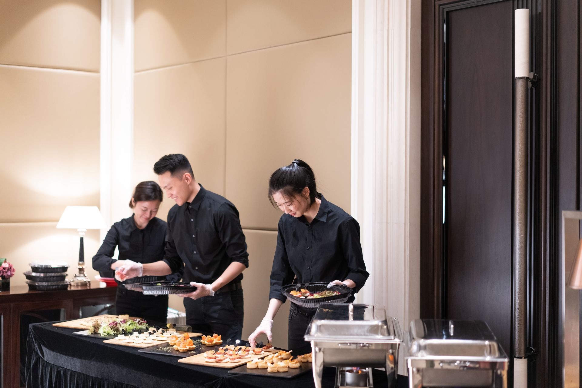 Three people in black uniforms setting up food on a buffet table.