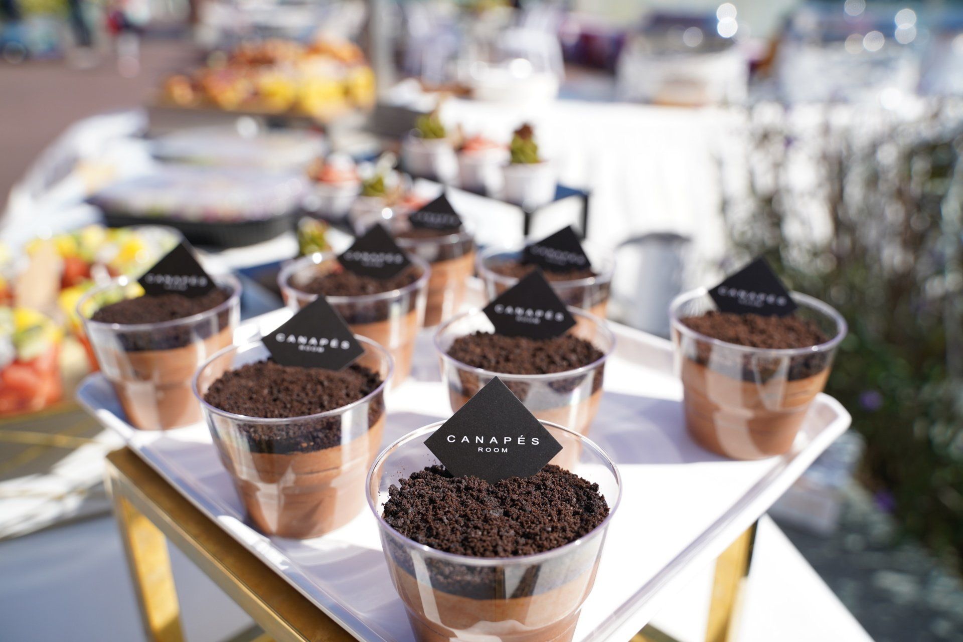Chocolate desserts in clear cups topped with cookies and logo labels on a white tray, outdoor setting.