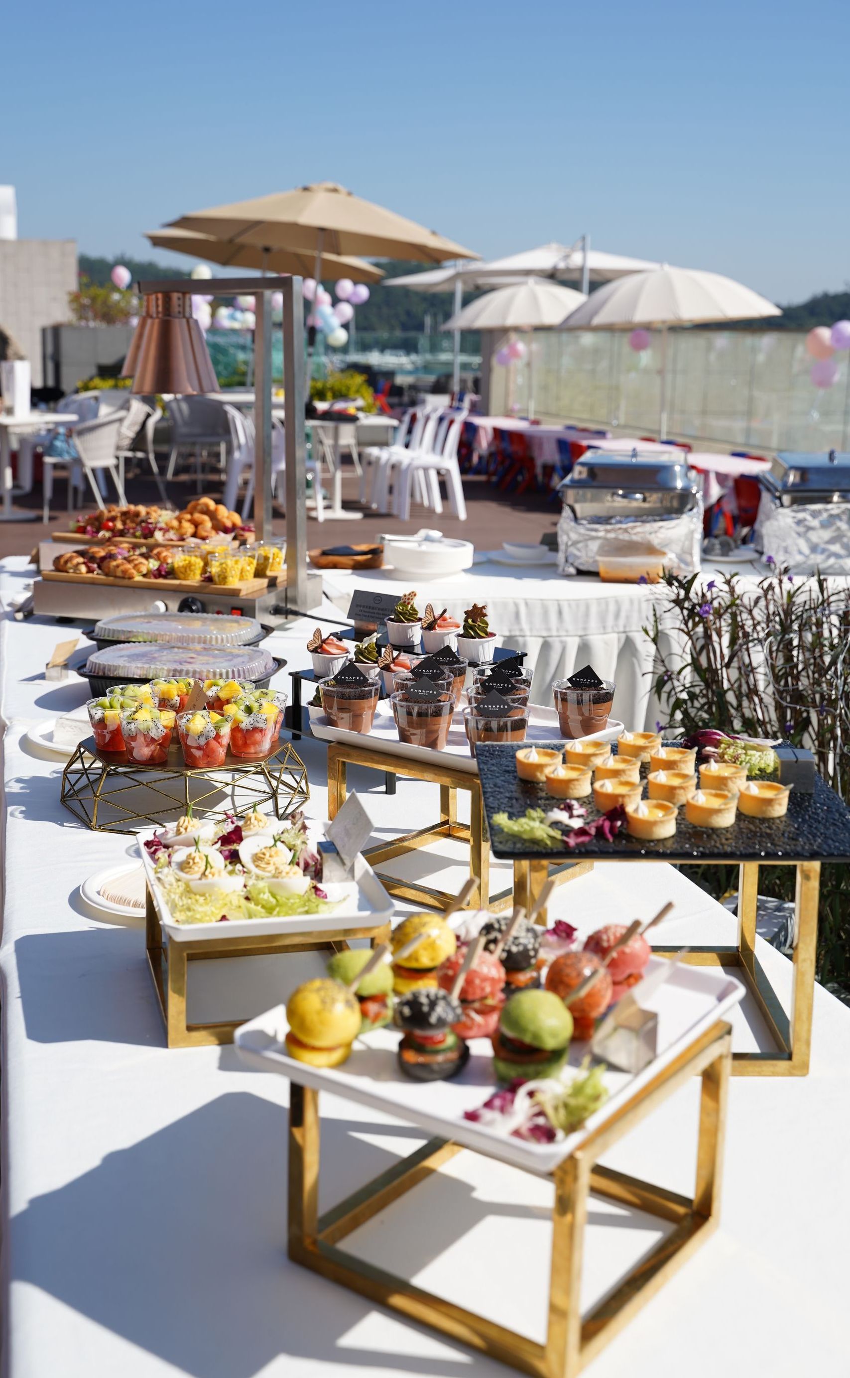 Buffet table with various foods set outdoors with umbrellas and chairs.
