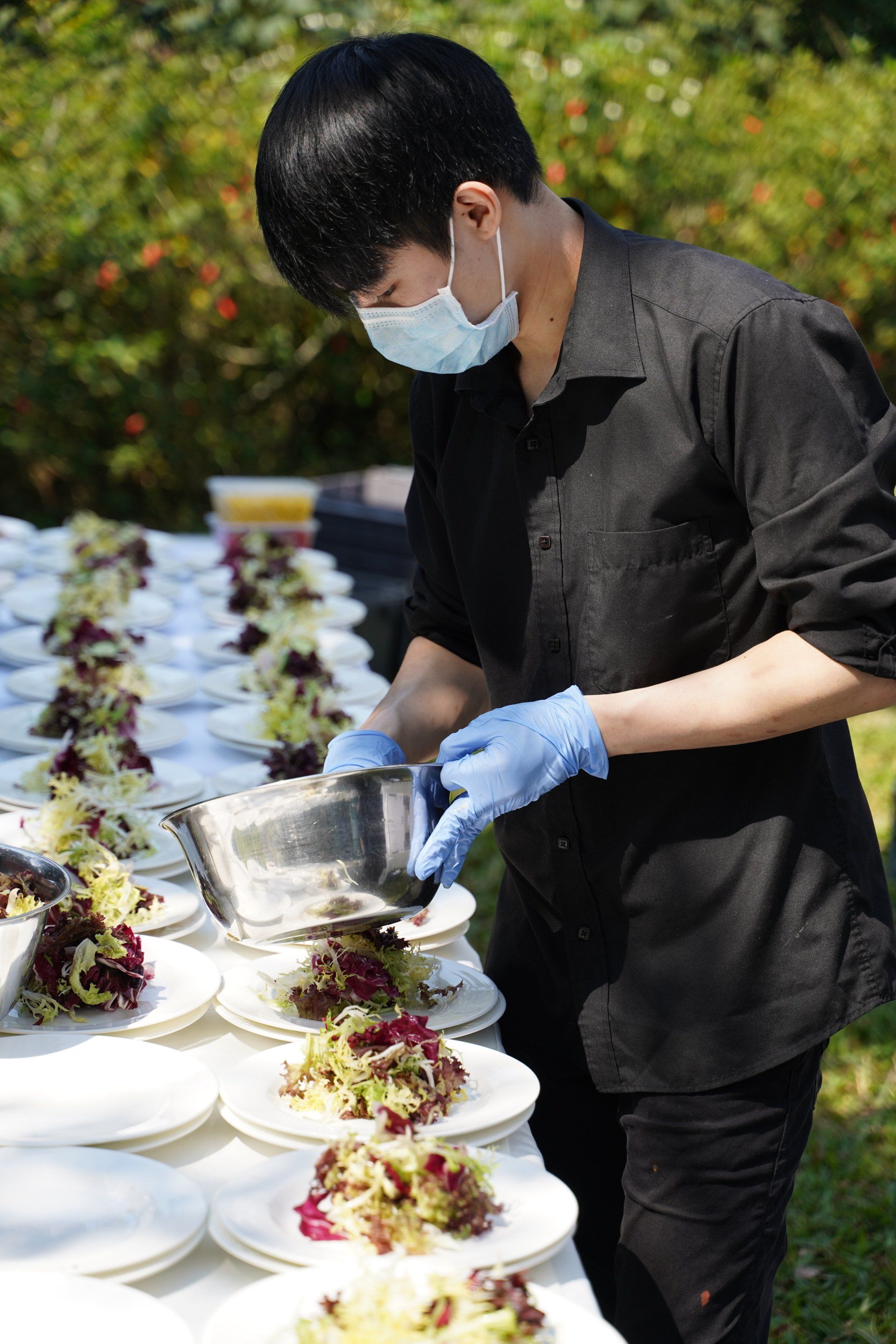 Person wearing mask and gloves preparing salads outdoors at a catered event.