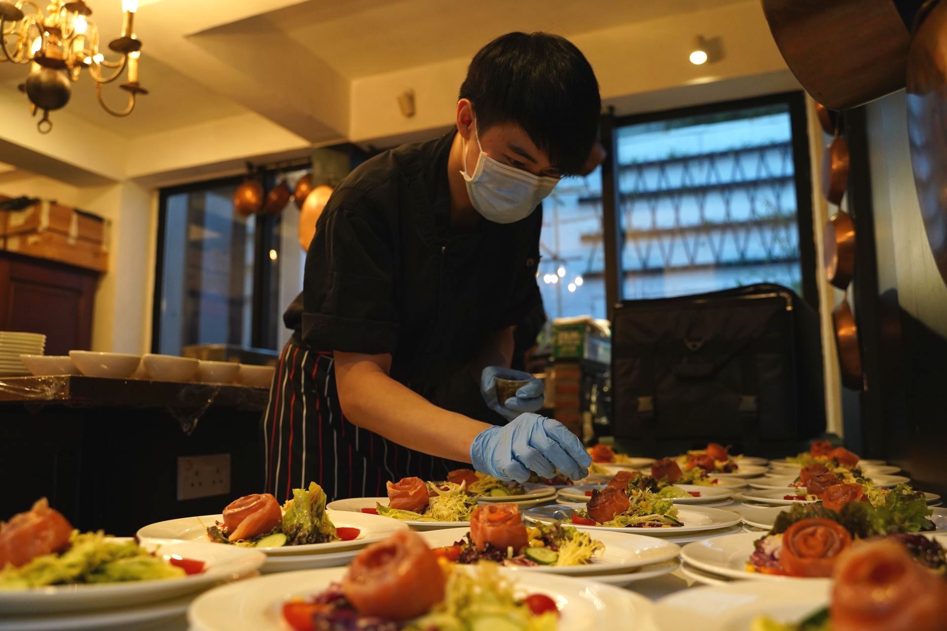 Chef plating dishes in a restaurant, wearing a mask and gloves. Plates with salmon and avocado are in the foreground.