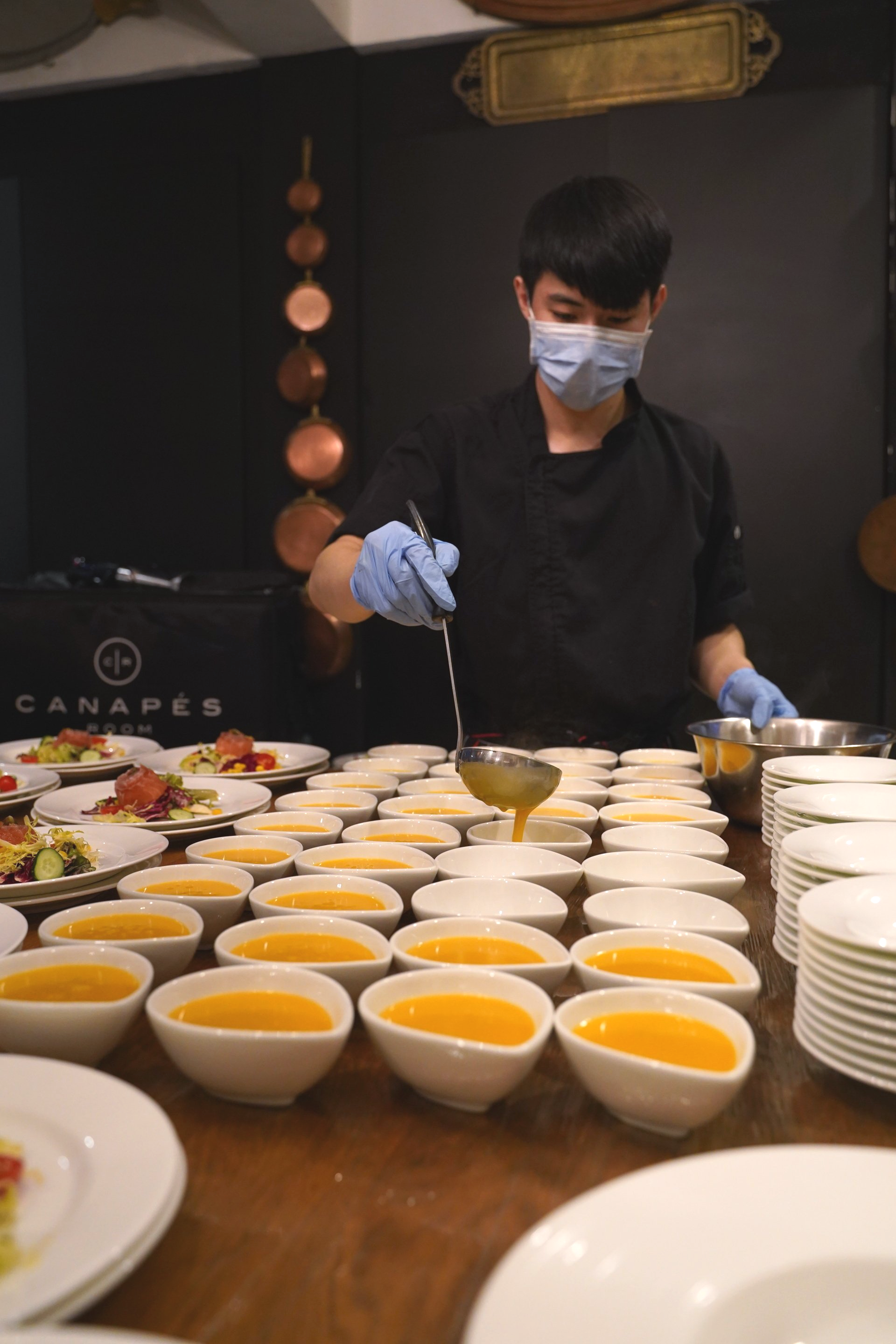 Chef wearing mask and gloves pours sauce into bowls on a table set for service.