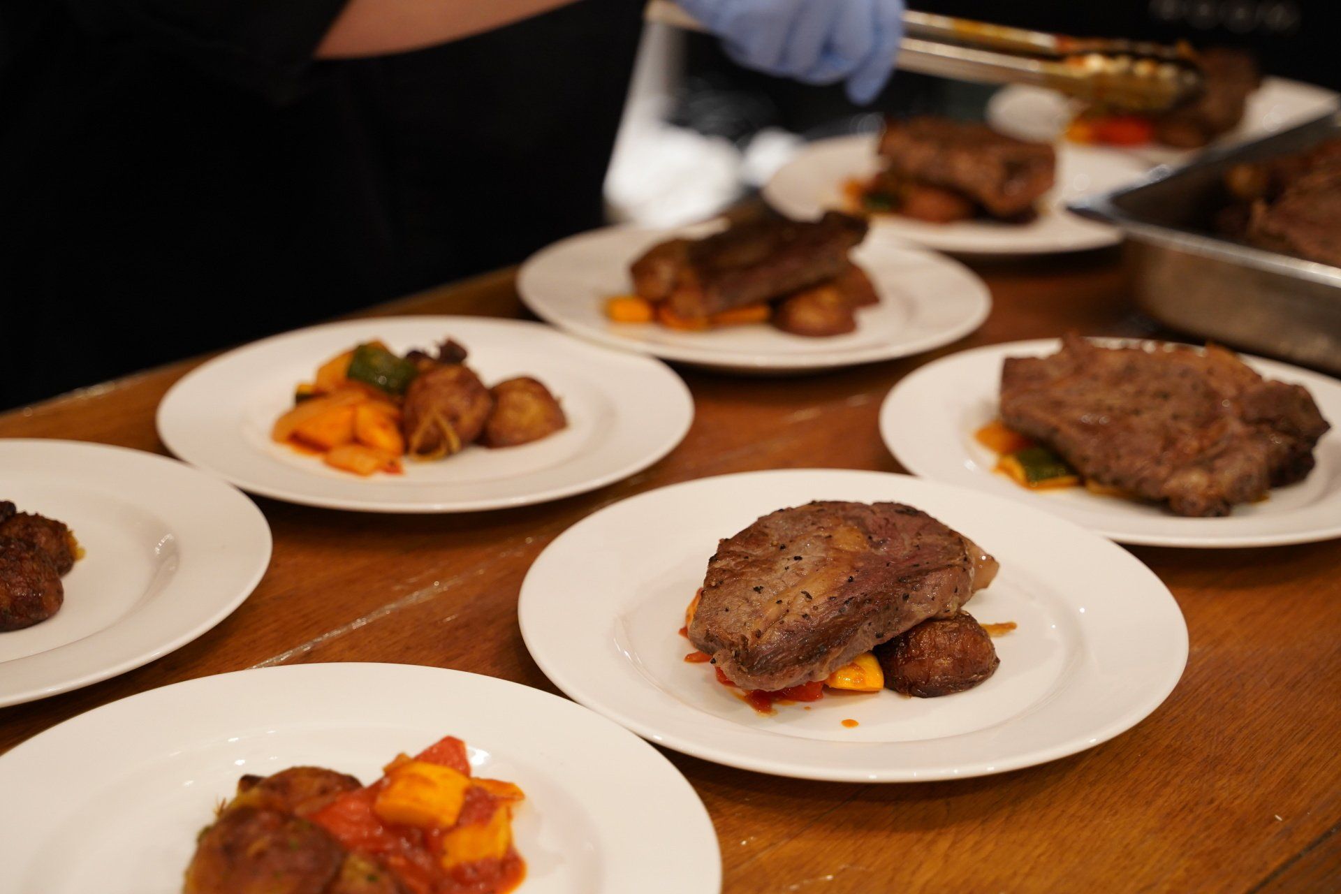 Plates of steak and roasted vegetables being prepared; gloved hands use tongs to serve.