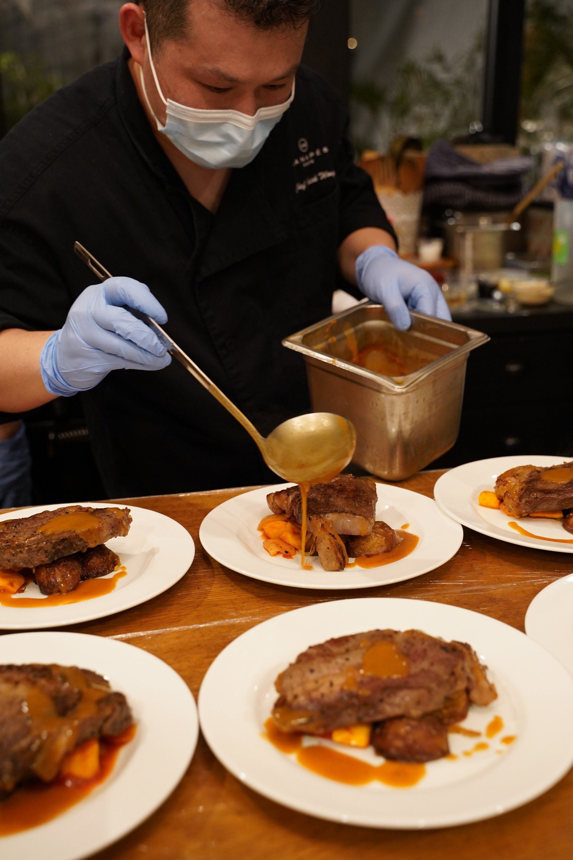Chef in mask and gloves plates food, pouring sauce over meat and vegetables on white plates.