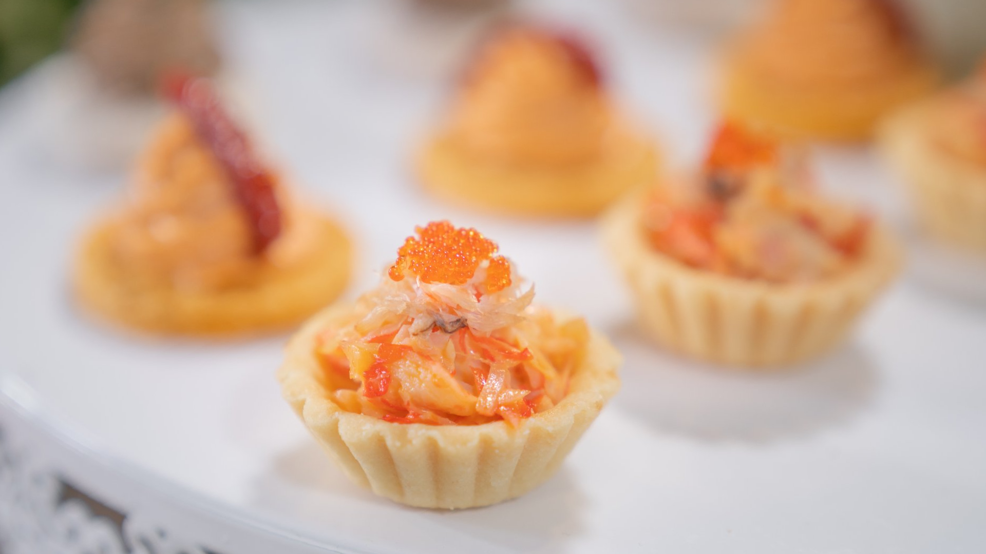 Mini savory tarts filled with orange seafood salad, topped with caviar, on a white tray.
