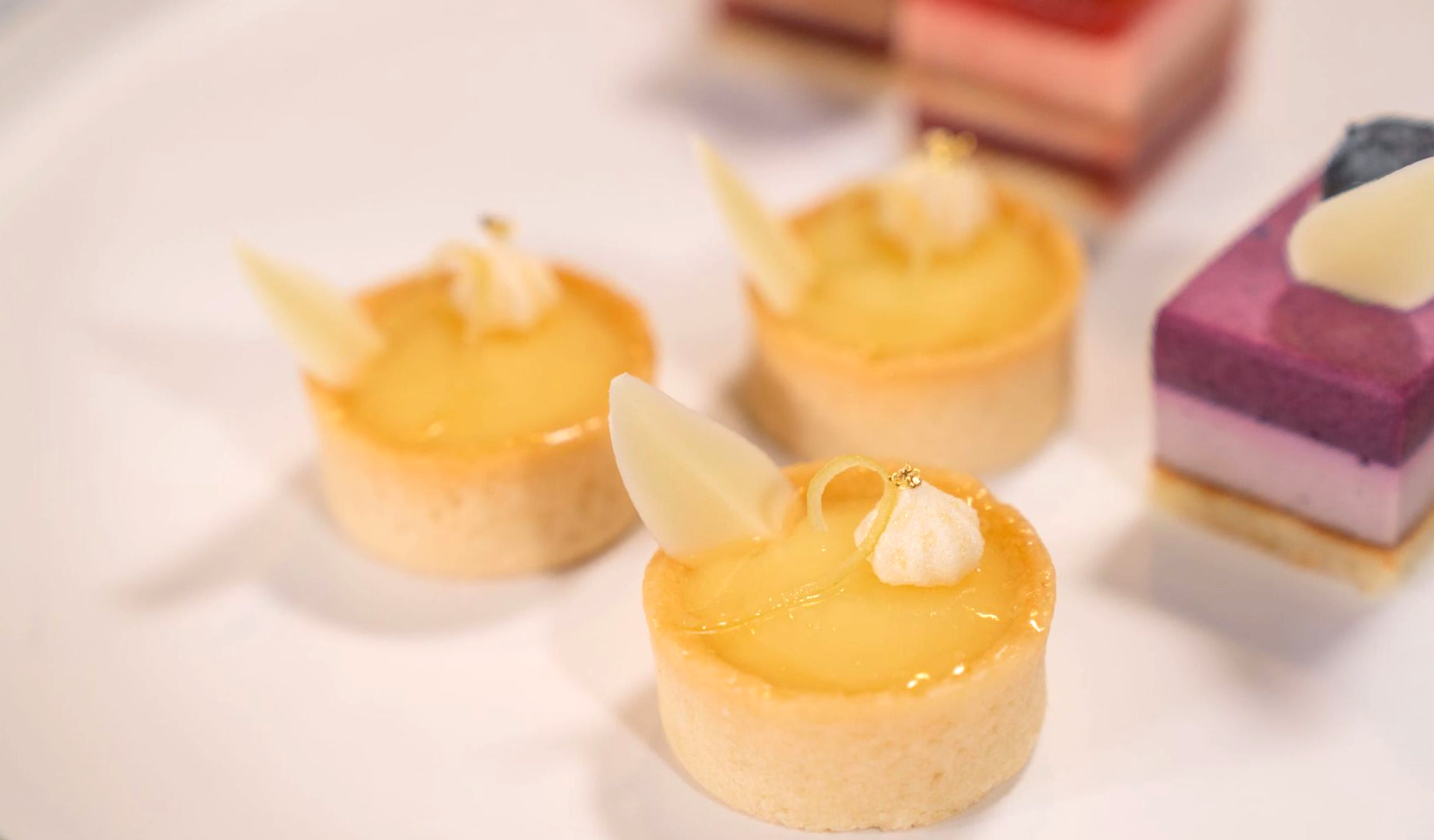 Mini lemon tarts with white chocolate garnish on a white plate, with layered desserts.