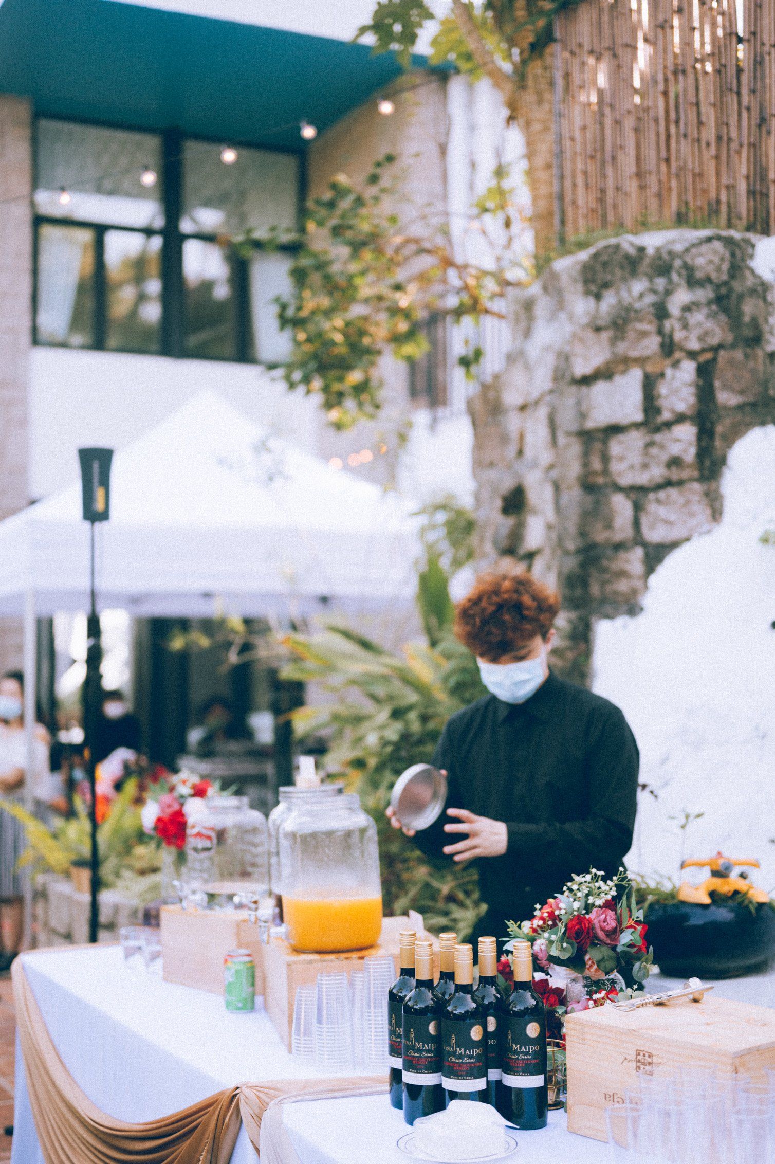 Bartender in mask prepares drinks at outdoor bar.
