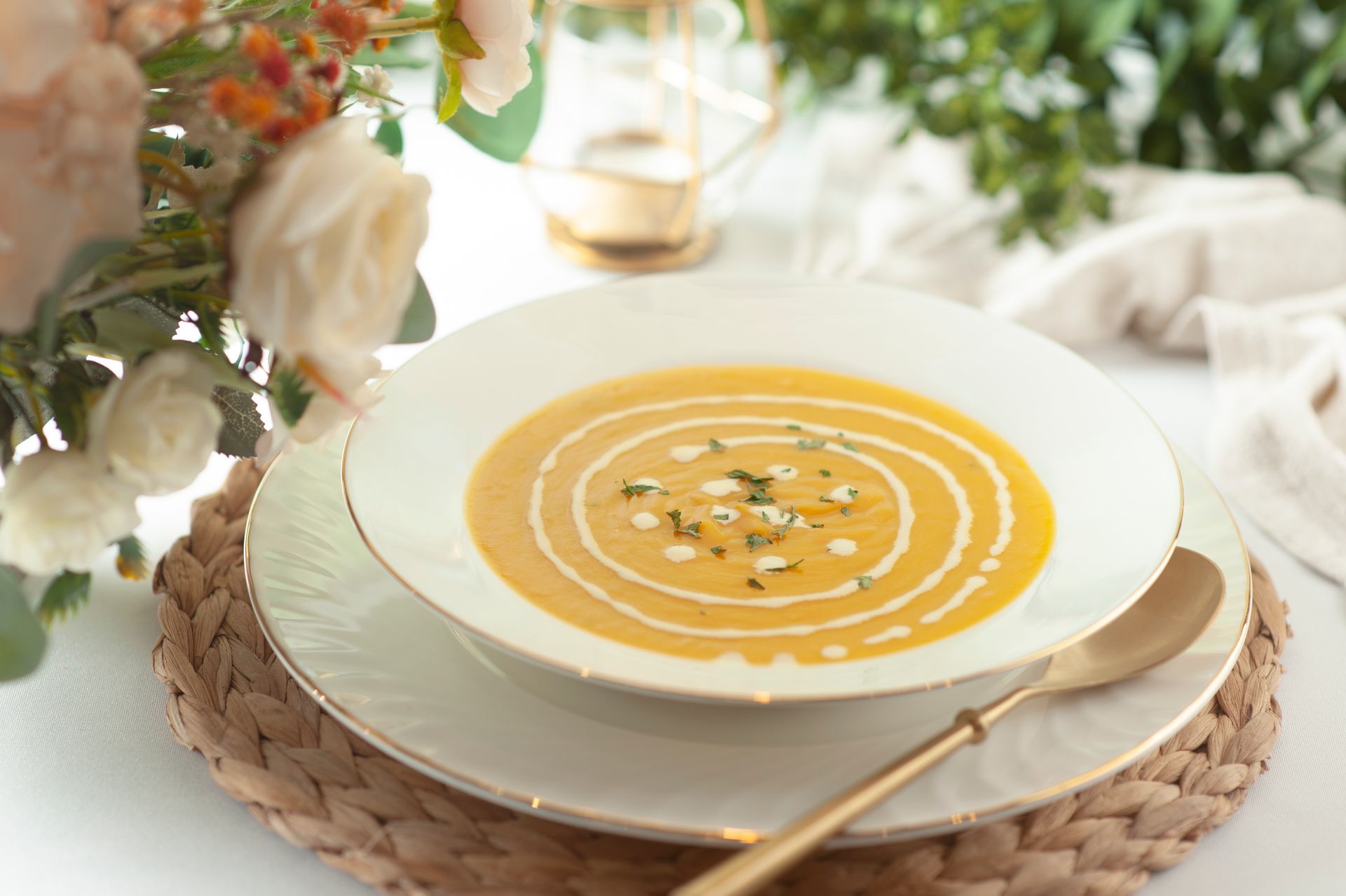 Creamy orange soup in white bowl with swirl, gold spoon, and floral arrangement.
