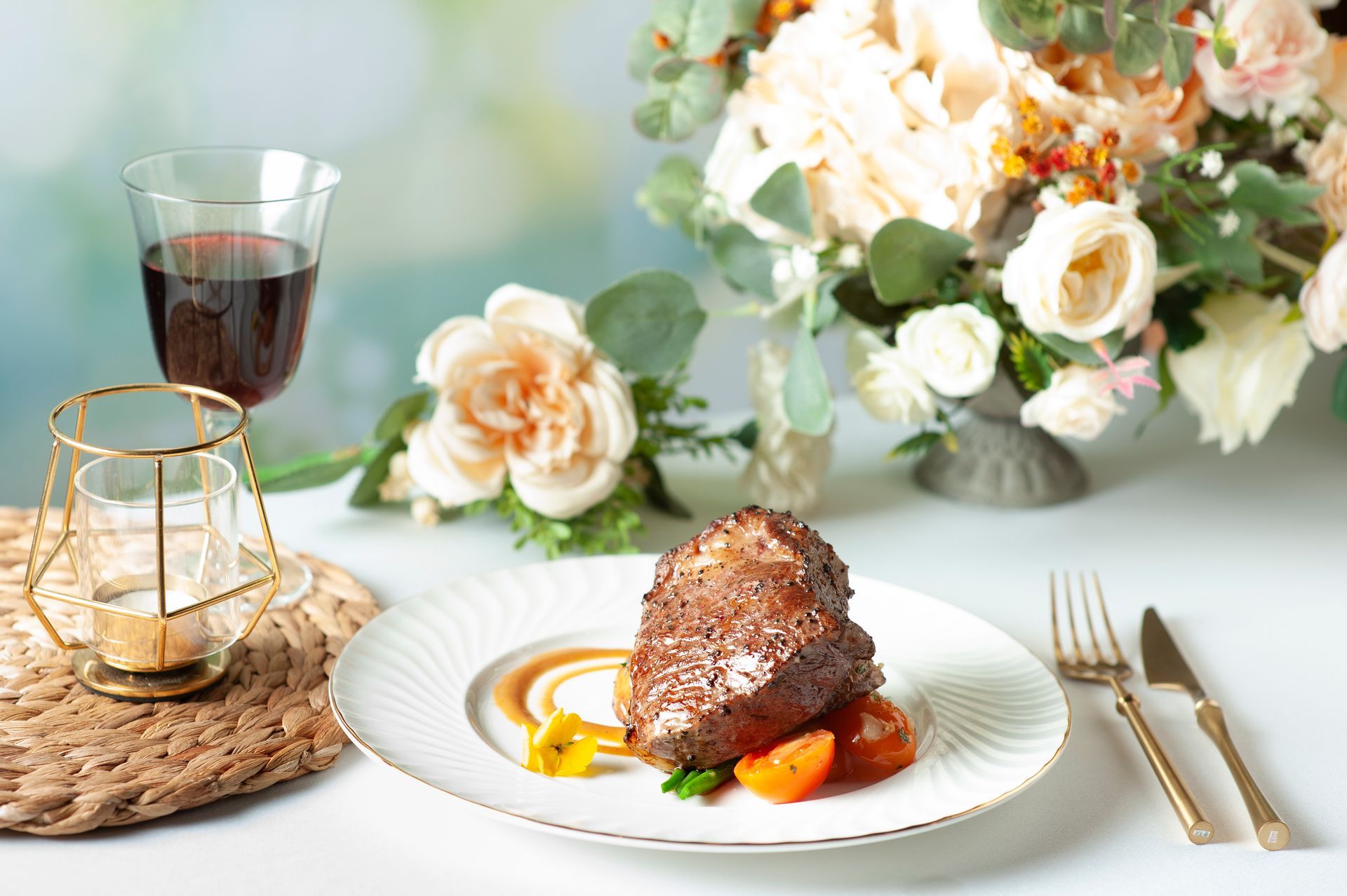 Steak on a white plate with sauce and tomato; wine glass, flowers, and gold candle holder on a table.