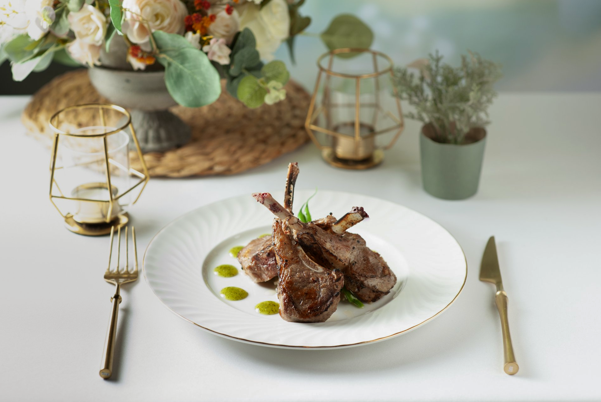 Lamb chops on a white plate with gold rim, fork, knife, and floral decor on a white table.