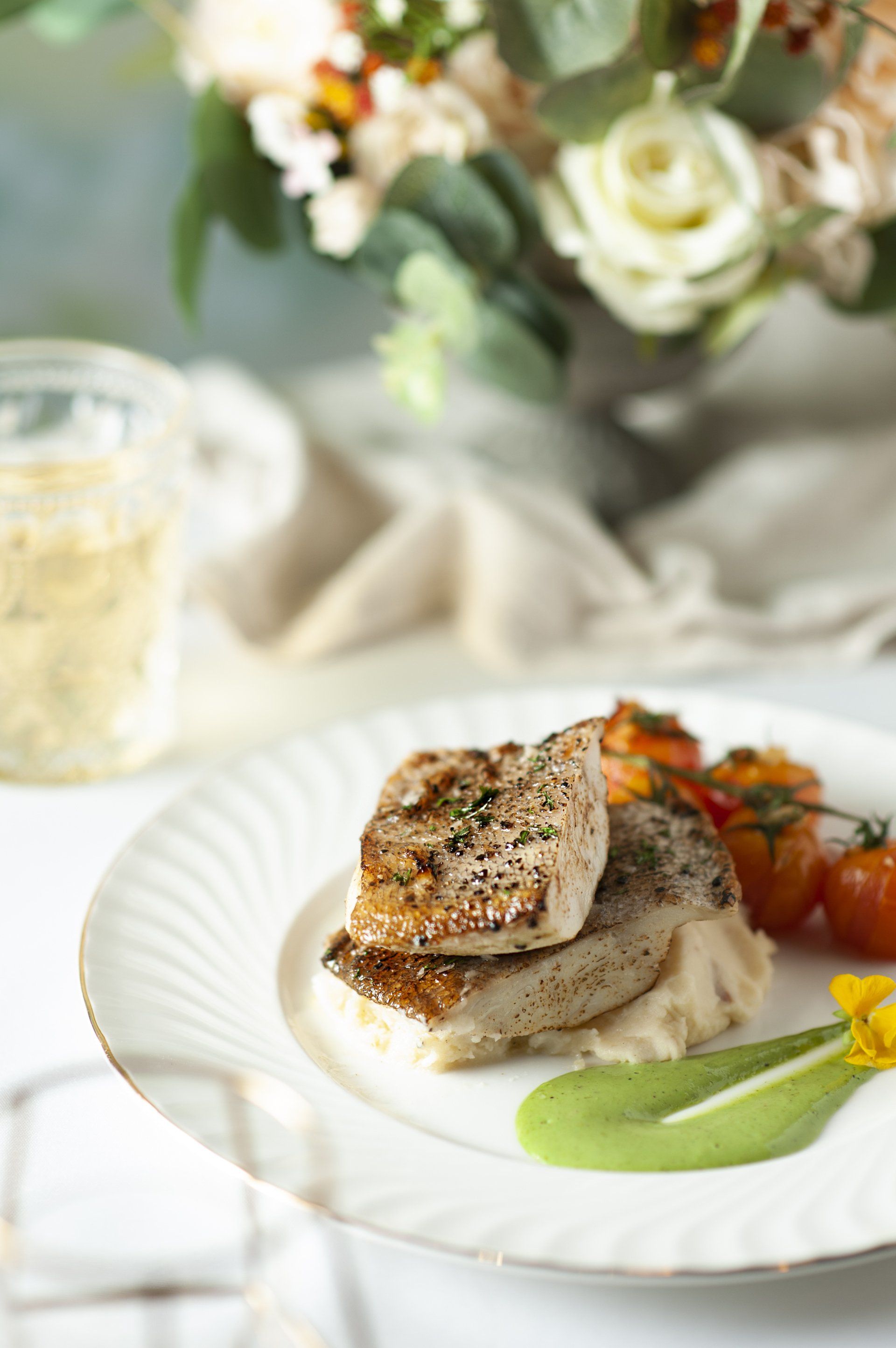 Grilled fish dish with tomatoes and a green sauce on a white plate, flowers in background.