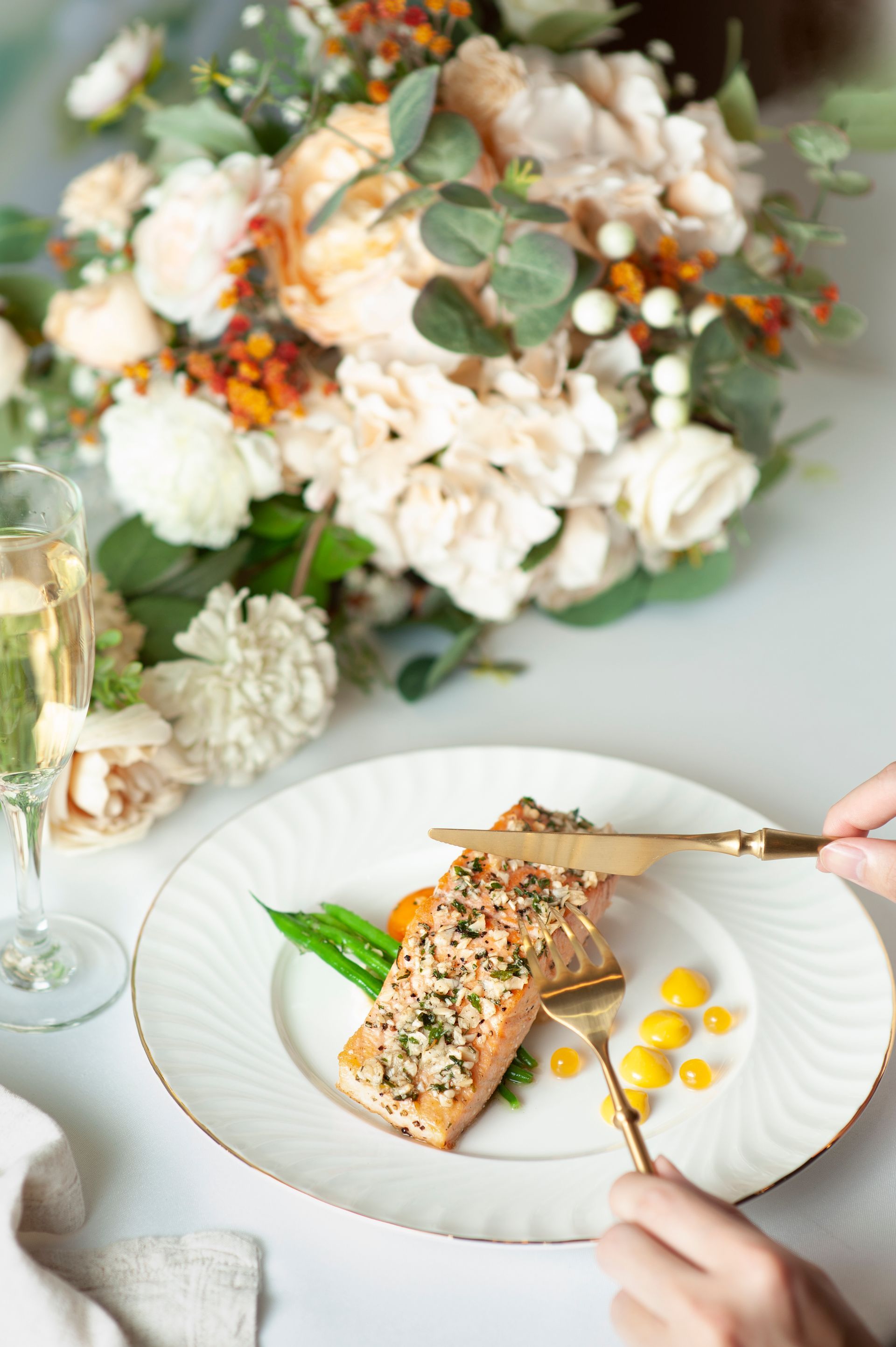Salmon dish on a white plate with gold cutlery. Bouquet of flowers in background.