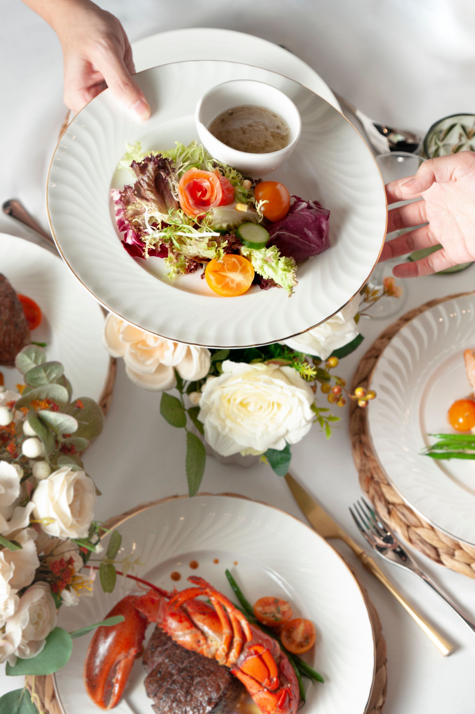 Elegant table setting with salad and lobster dishes, held by hands. White plates with gold trim.