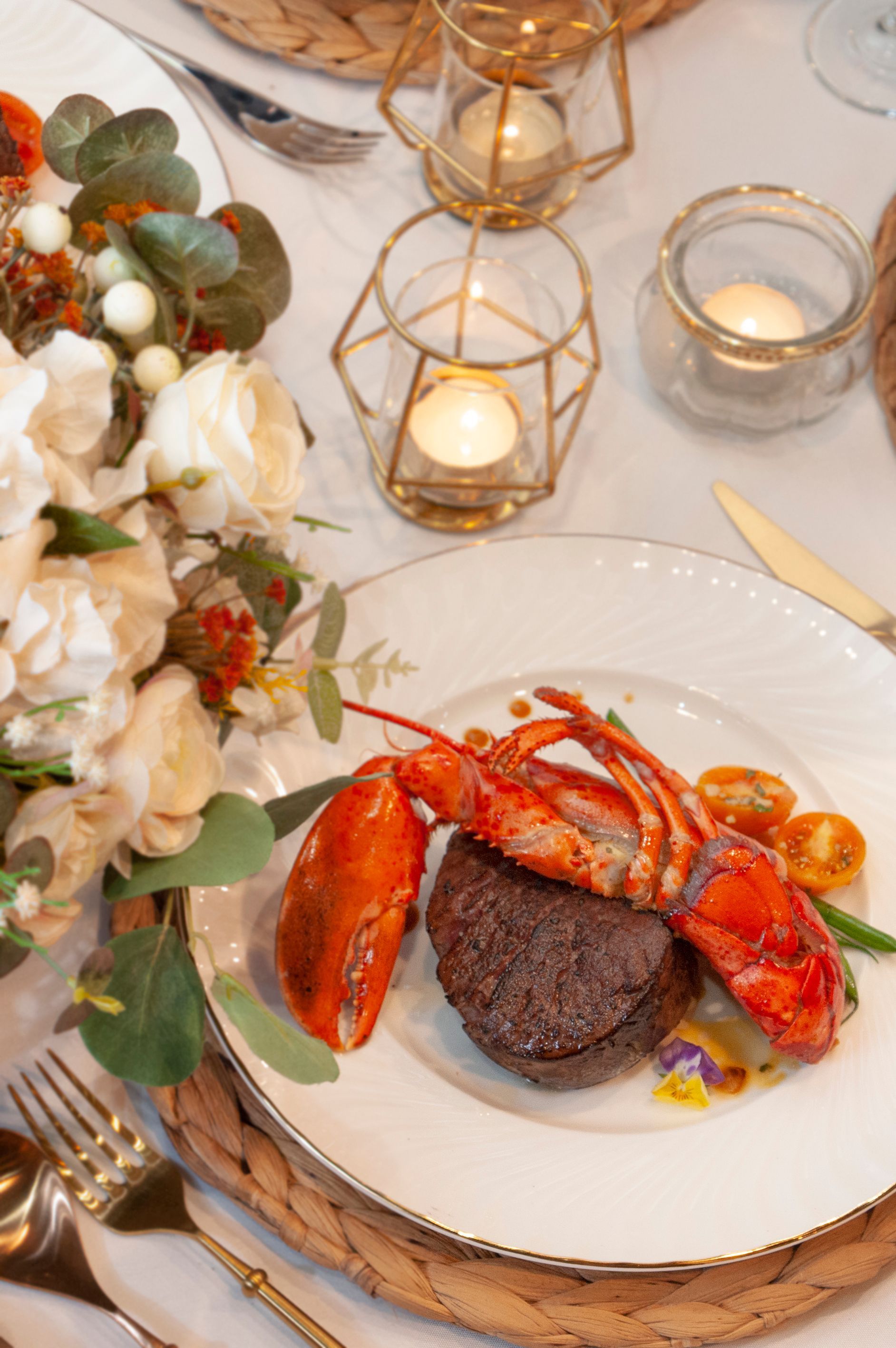 Lobster tail and steak on a plate at a formal dinner table, with flowers and candles.