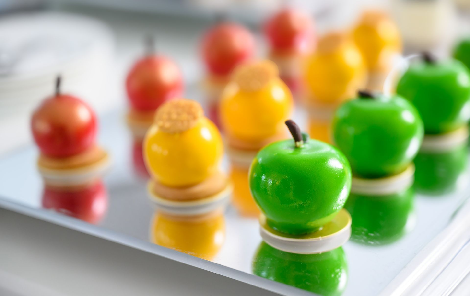 Colorful, apple-shaped desserts on a mirrored tray; red, yellow, and green, atop small, layered bases.