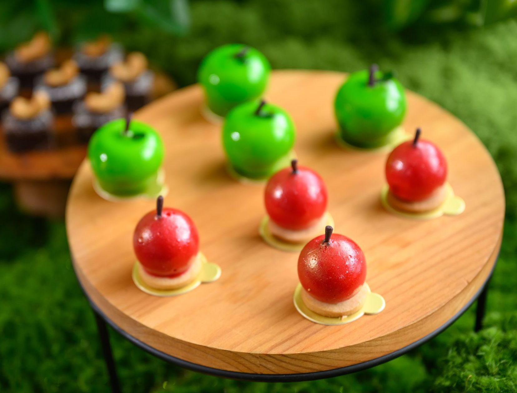 Wooden tray with red and green apple-shaped treats on a mossy background.