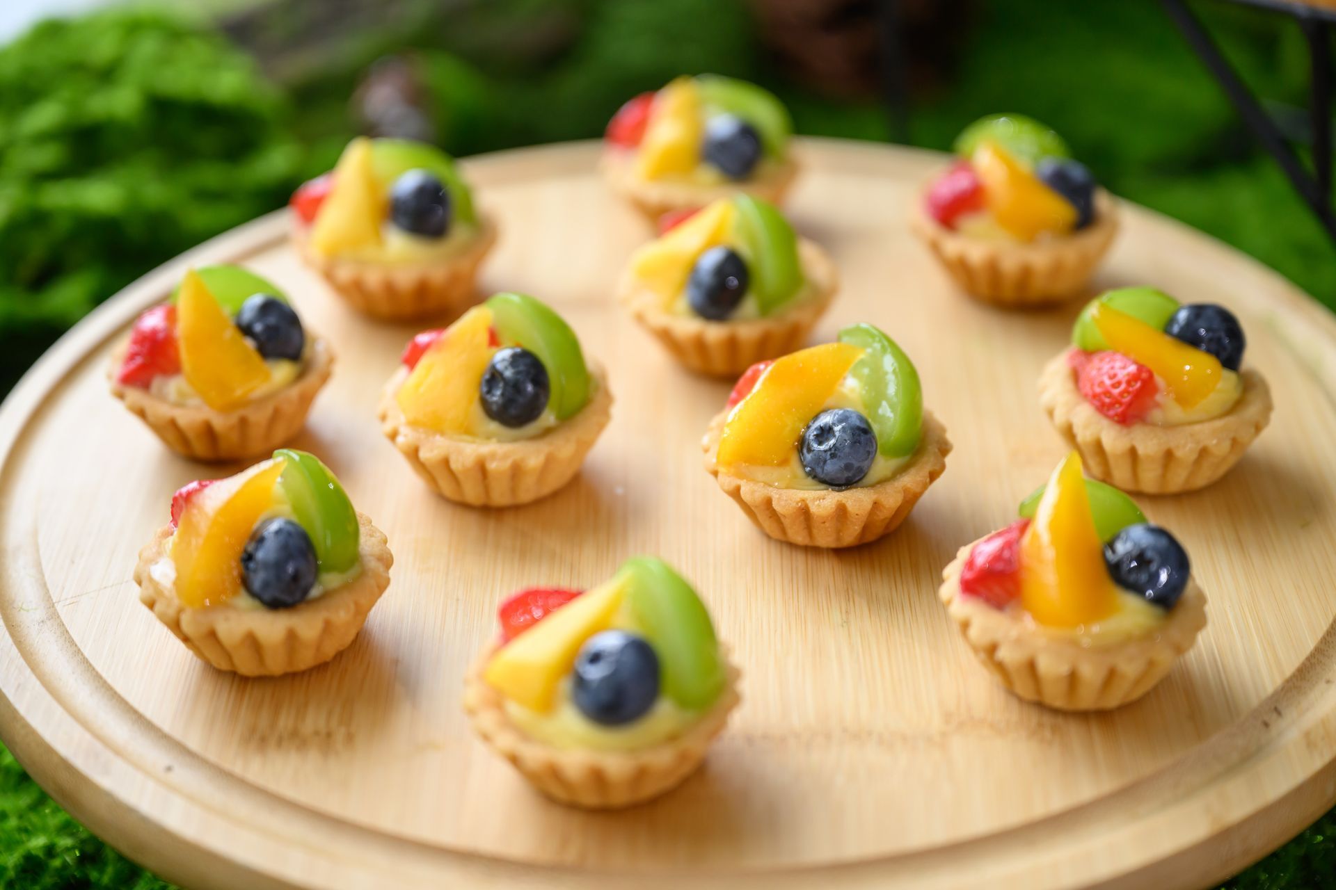 Mini fruit tarts on a wooden tray, each with a crust filled with custard and topped with fruit.