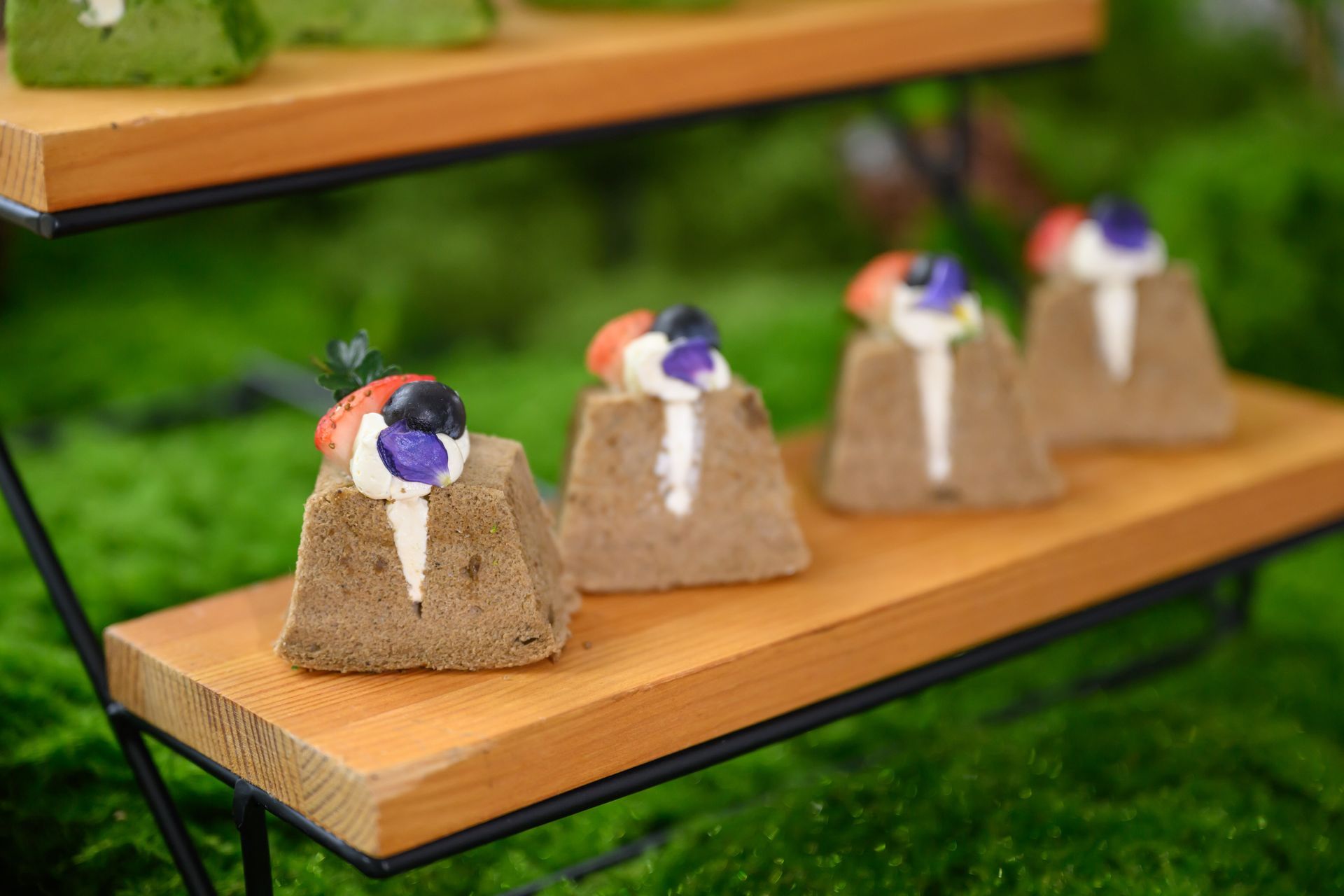 Cakes on wooden tiered stand. Brown sponge cakes topped with berries and cream. Green moss background.