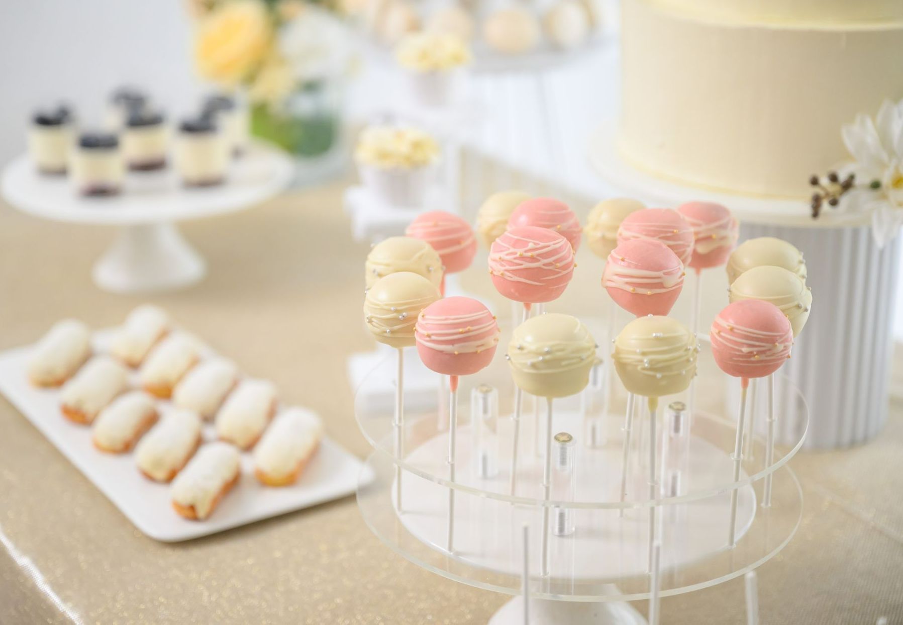 Dessert table with pink and white cake pops, eclairs, and a tiered cake.