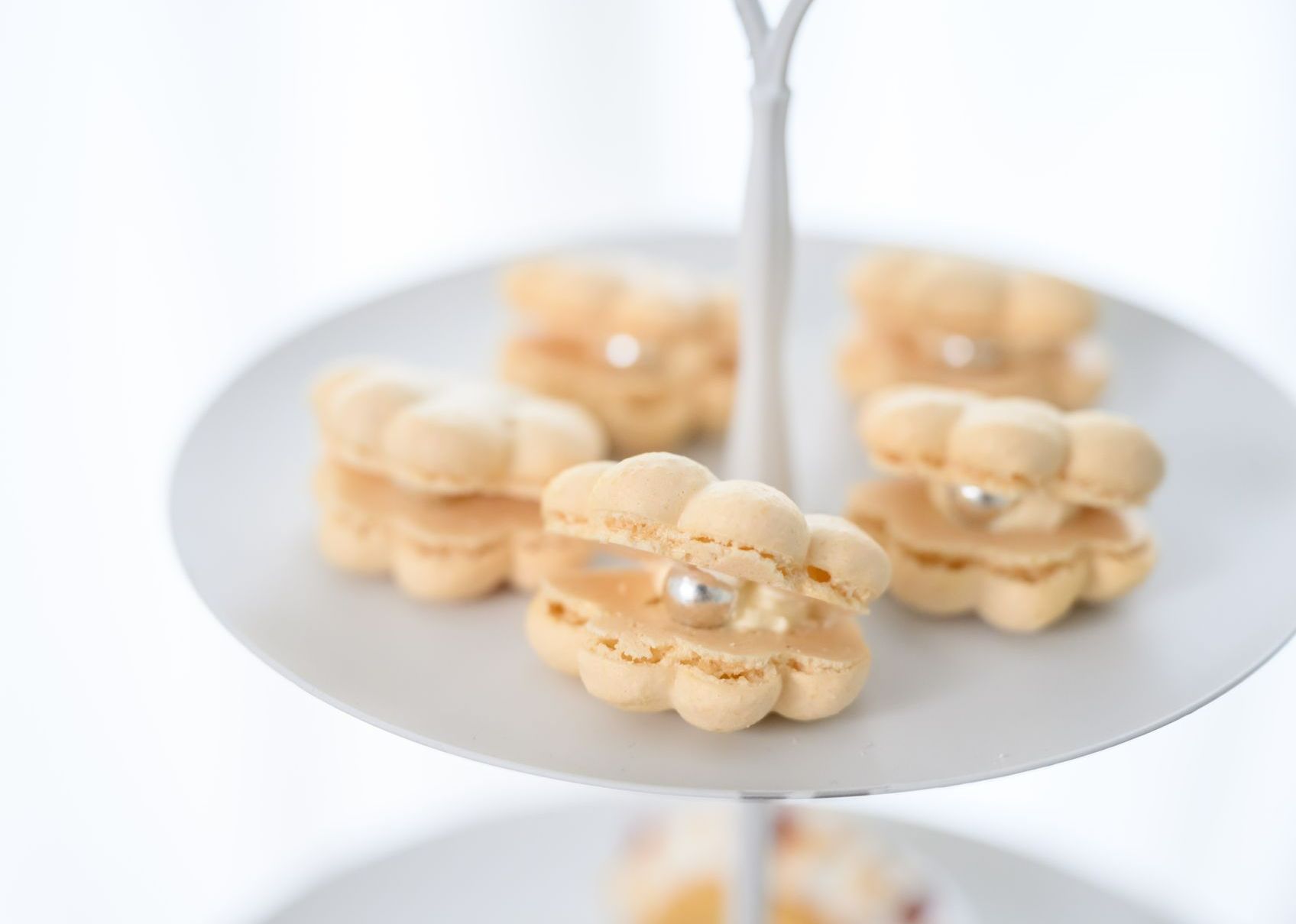 Flower-shaped macarons on a tiered white serving tray, with cream filling and a silver sugar pearl.