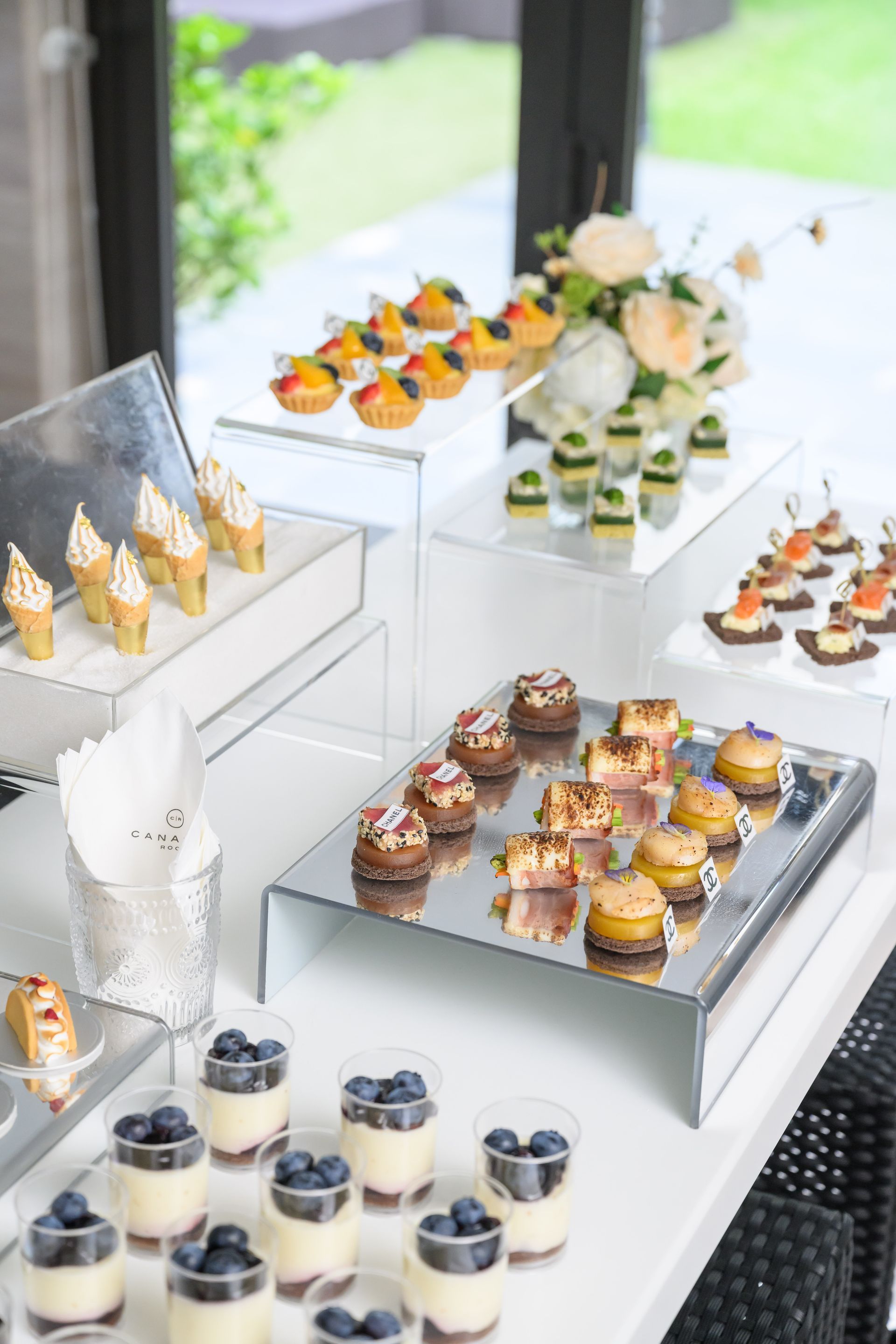 Dessert buffet on a white table with mini pastries and parfaits; floral arrangement in background.