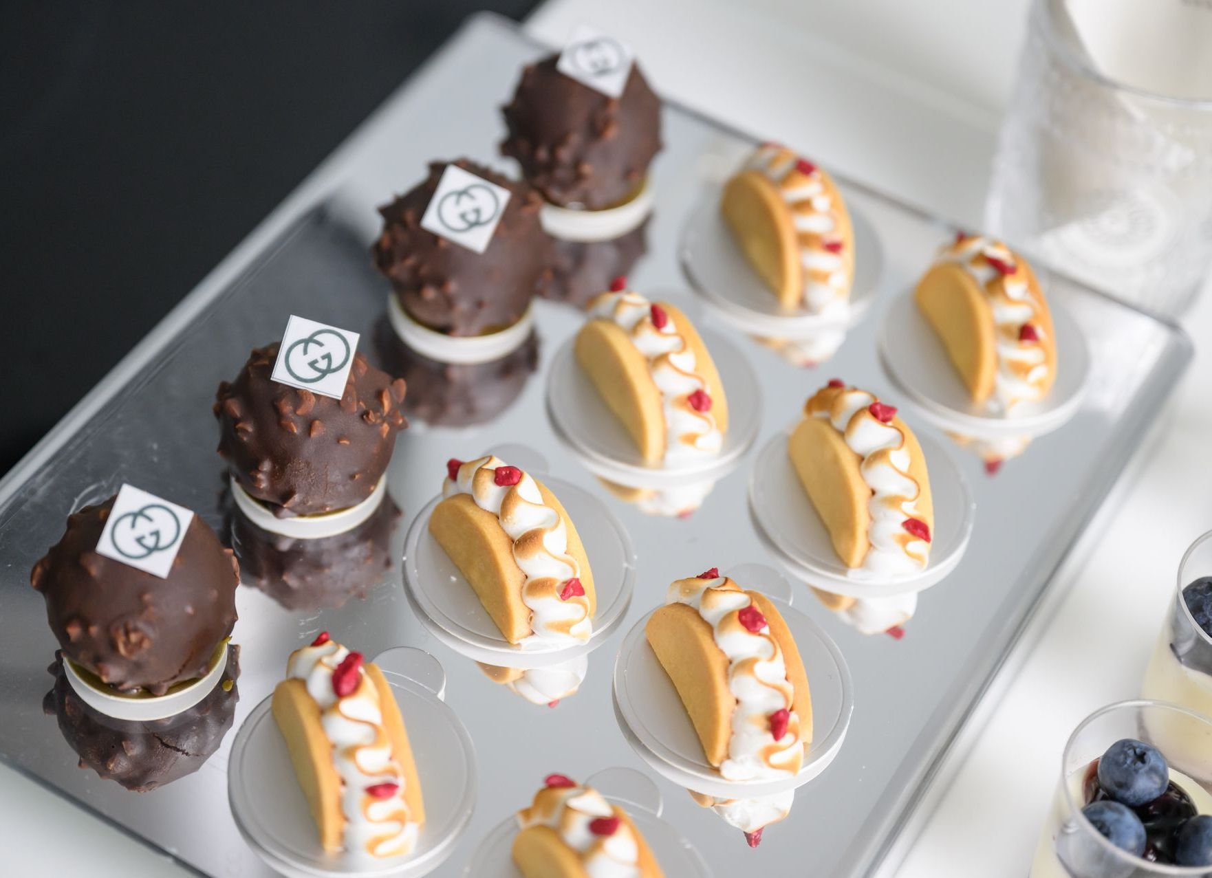 Tray of miniature desserts: chocolate spheres with logo, taco-shaped sweets with topping, on glass dishes.