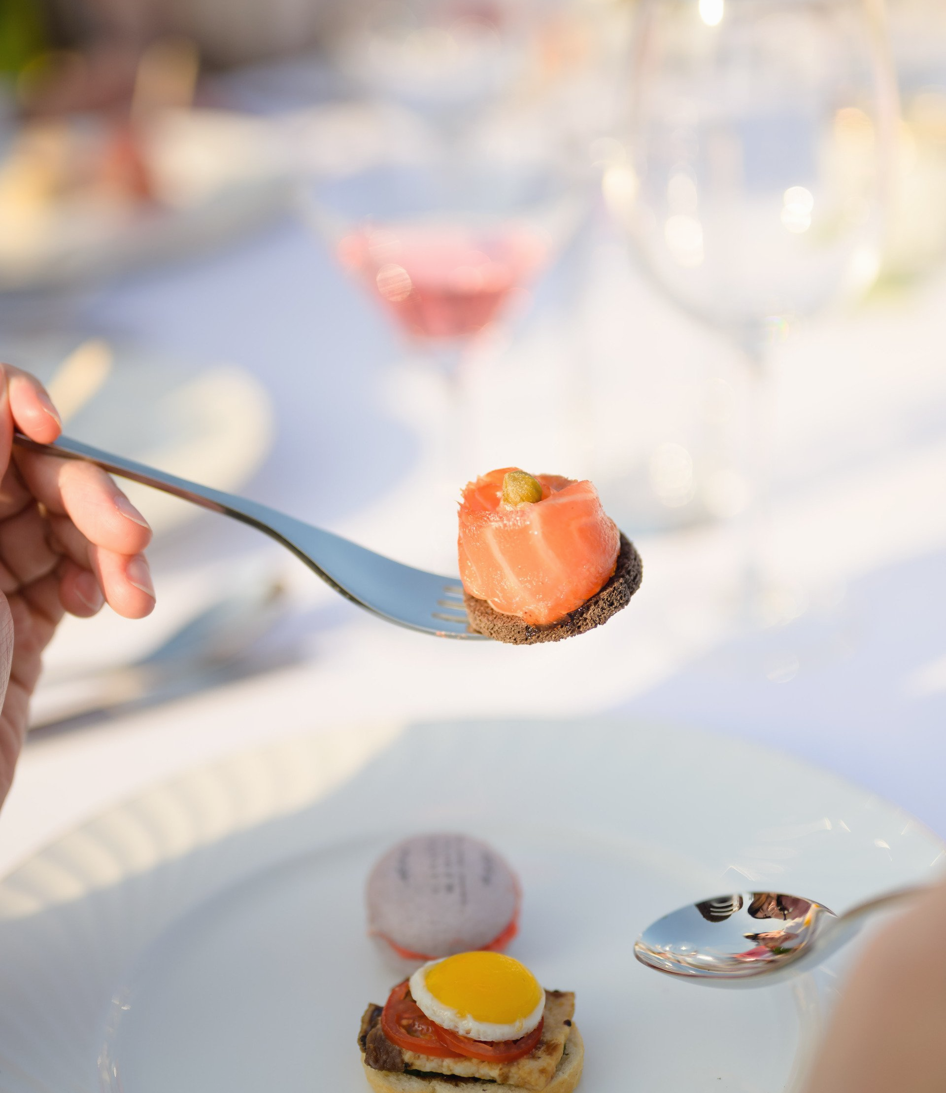 Person holding a spoon with a salmon bite, served with other appetizers on a white plate. Cocktail glass in background.