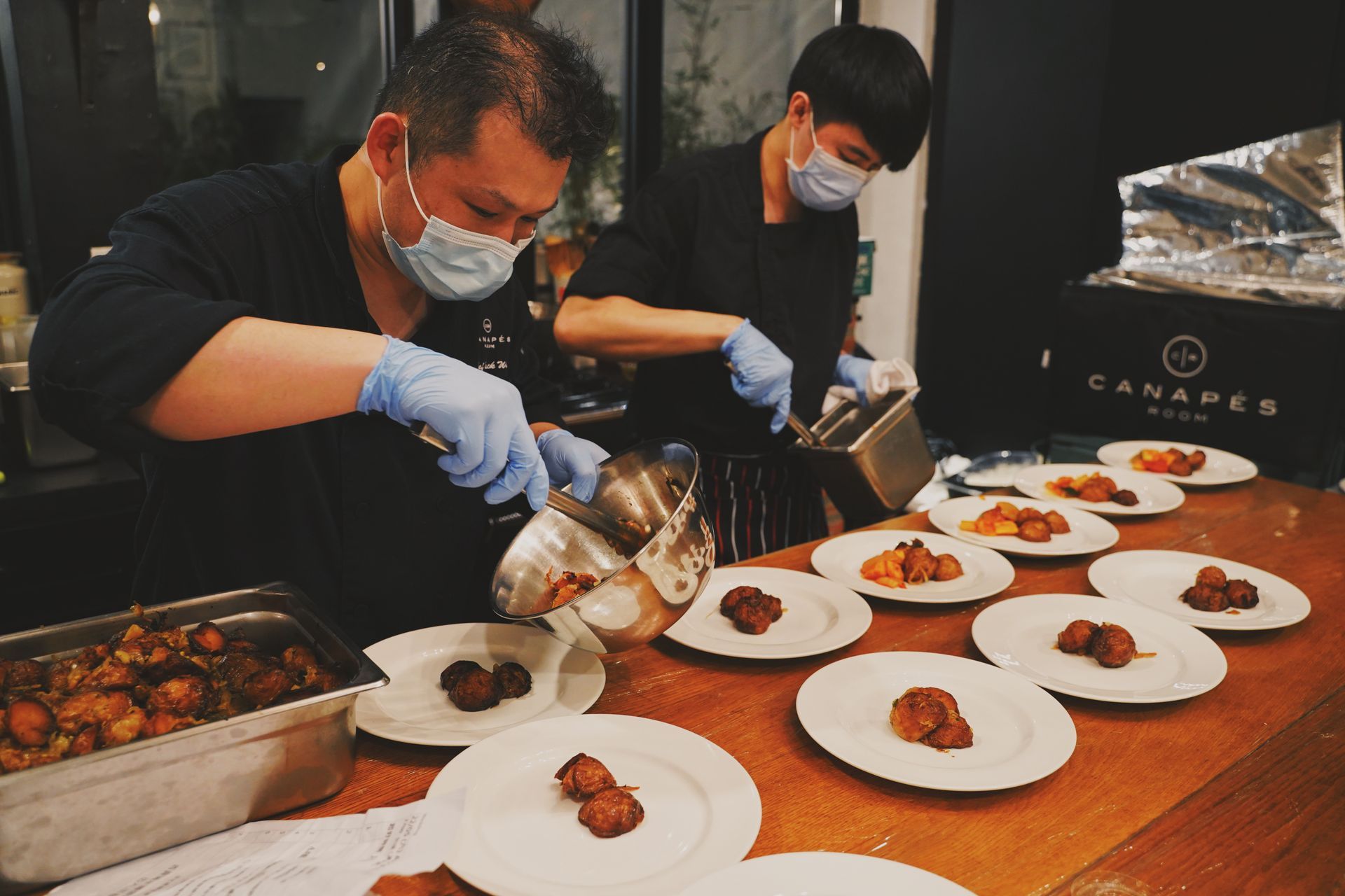 Two chefs plating food, wearing masks and gloves, at a restaurant.