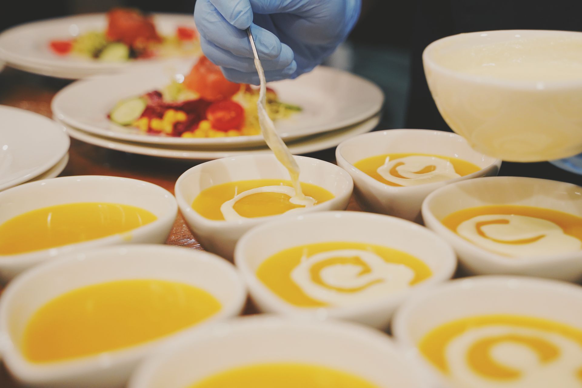 Bowls of yellow soup being swirled with white cream by a gloved hand in a kitchen.