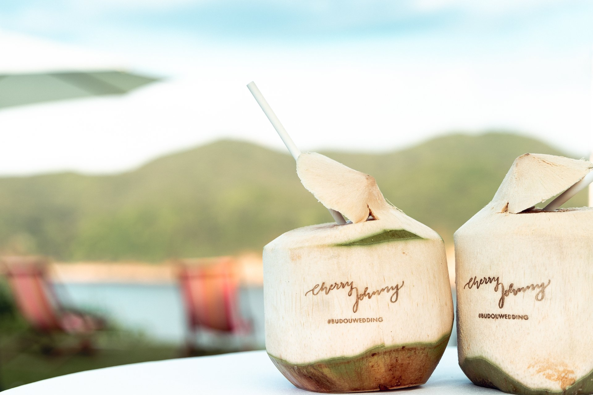 Two coconuts with straws on a white table, mountains and chairs in background.