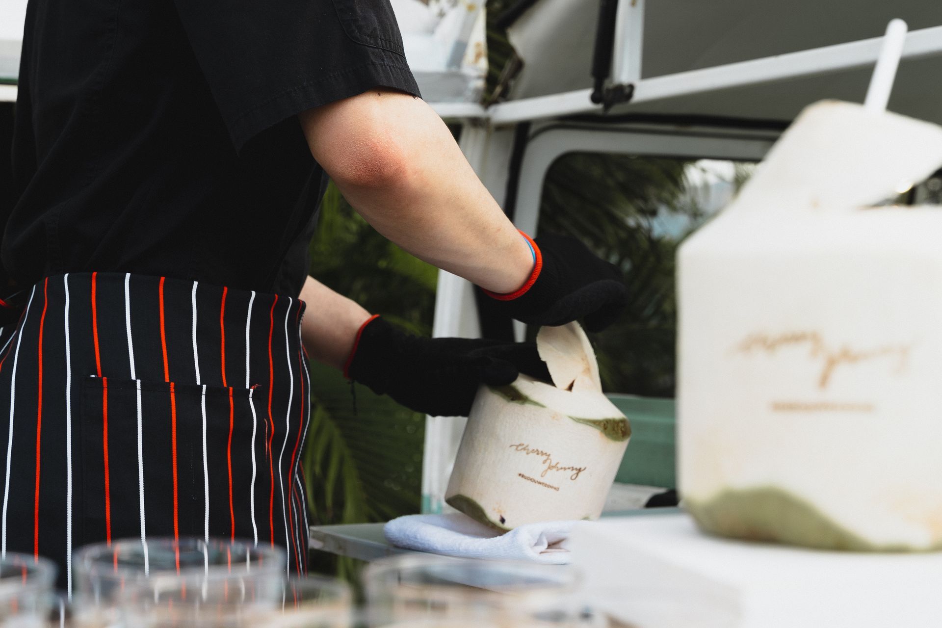 Person opening a coconut at a food stand, wearing black gloves and striped apron.
